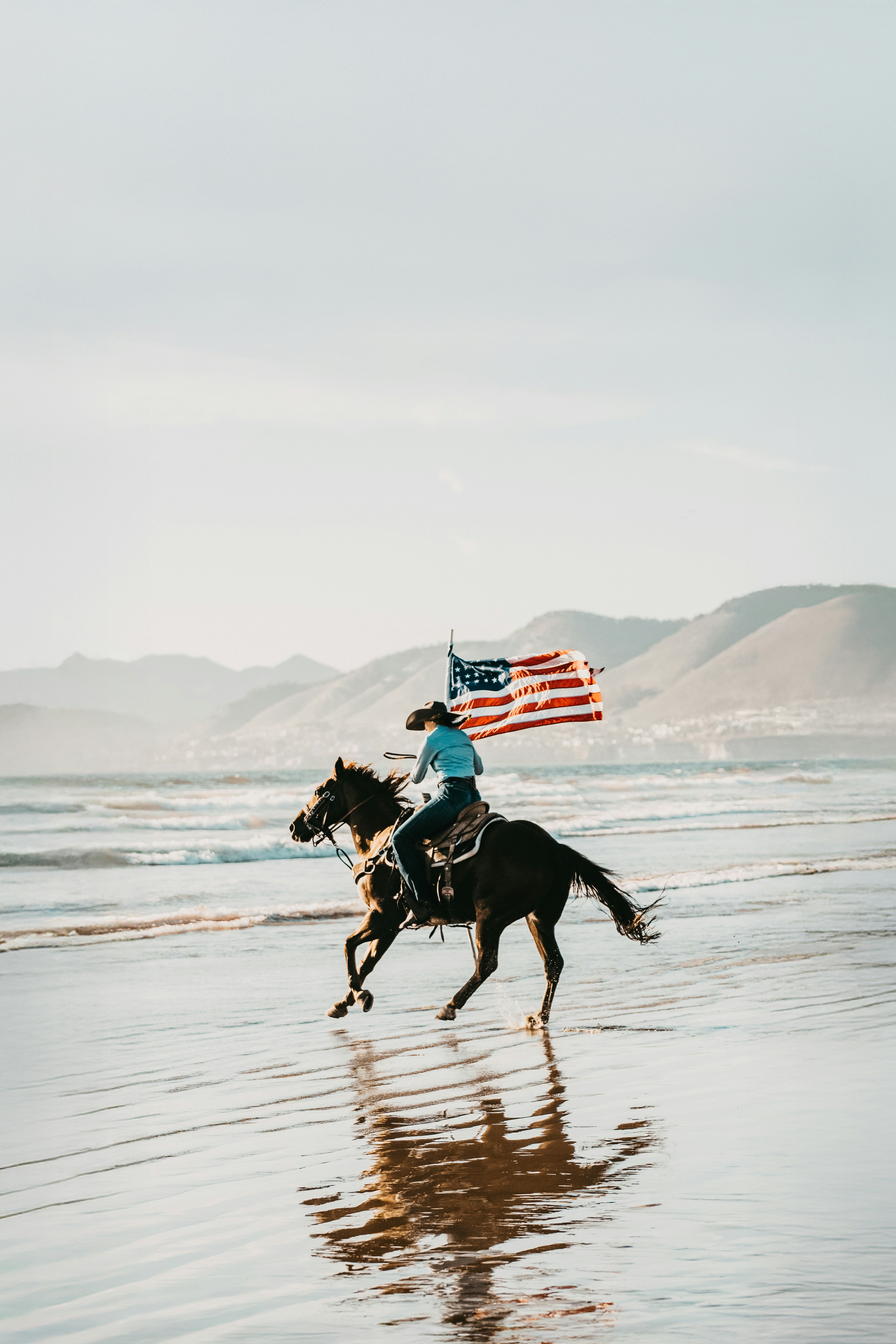 Cowgirl rides horse with american flag on beach.