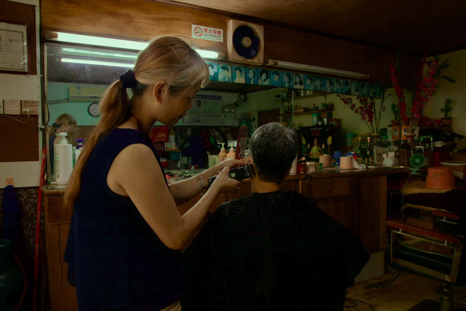 In a dimly lit room, a woman with long blonde hair is cutting another person's hair using a pair of scissors. The person being cut has their back to the camera and appears to be wearing a black shirt. Various bottles are scattered around the room, suggesting that this might be a barber shop or salon setting.