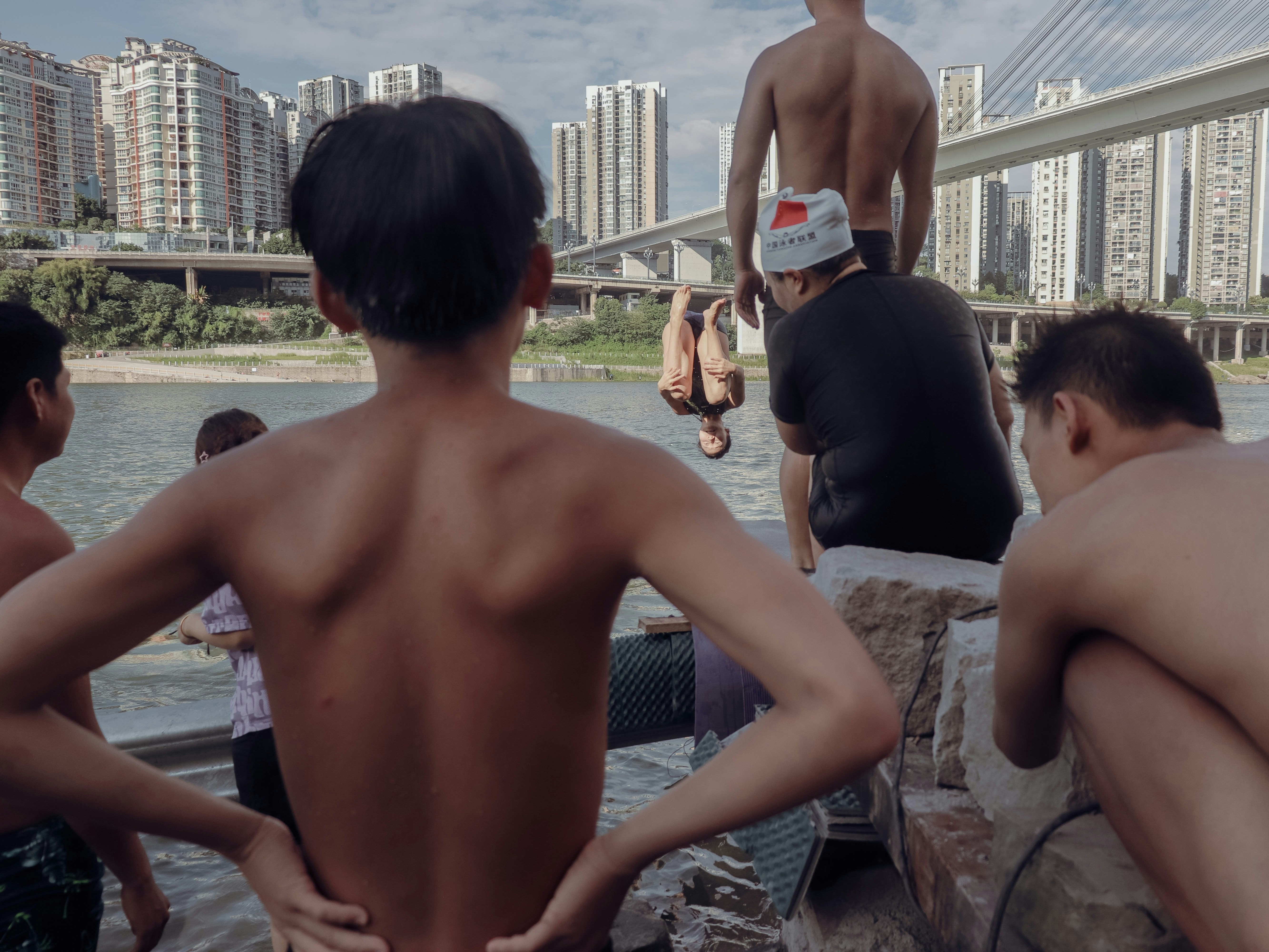 Group of shirtless men by the water with city buildings.
