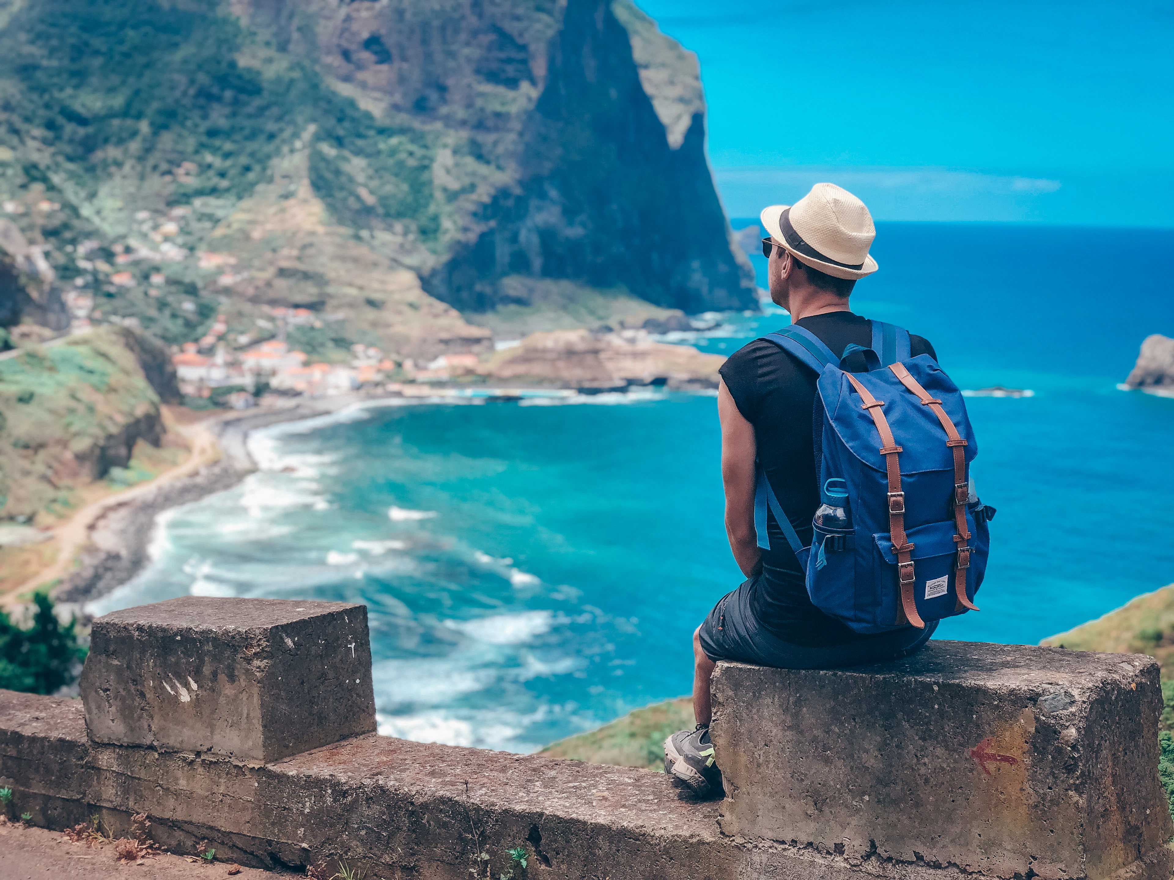 A traveller with a backpack sits on a stone wall above a coastal village and a turquoise bay