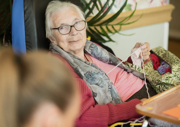 Elderly woman knitting in a wheelchair with a soft smile.