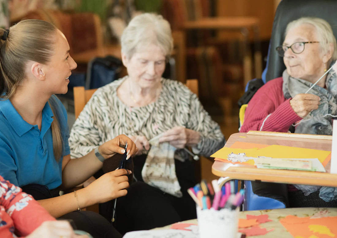A day program staff member in uniform sitting with two participants working on their knitting at a table in a community activity room