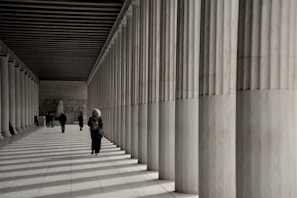 People walk along a colonnade with striped shadows.