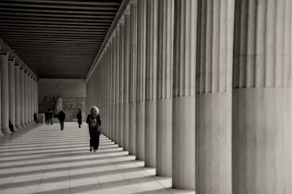 People walk along a colonnade with striped shadows.