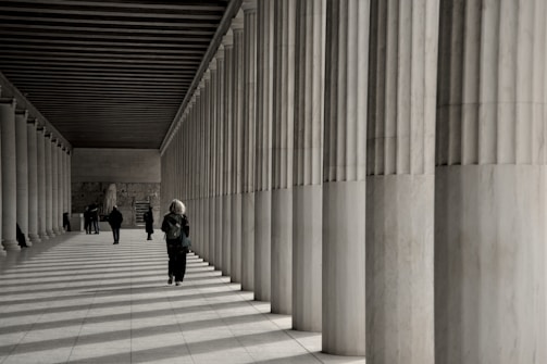 People walk along a colonnade with striped shadows.