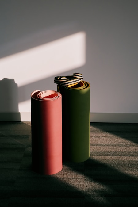 Two rolled yoga mats, one pink and one olive with a carry strap, standing against a white wall