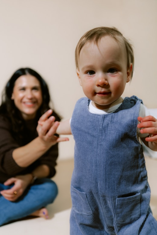 A baby in blue overalls with a woman behind.