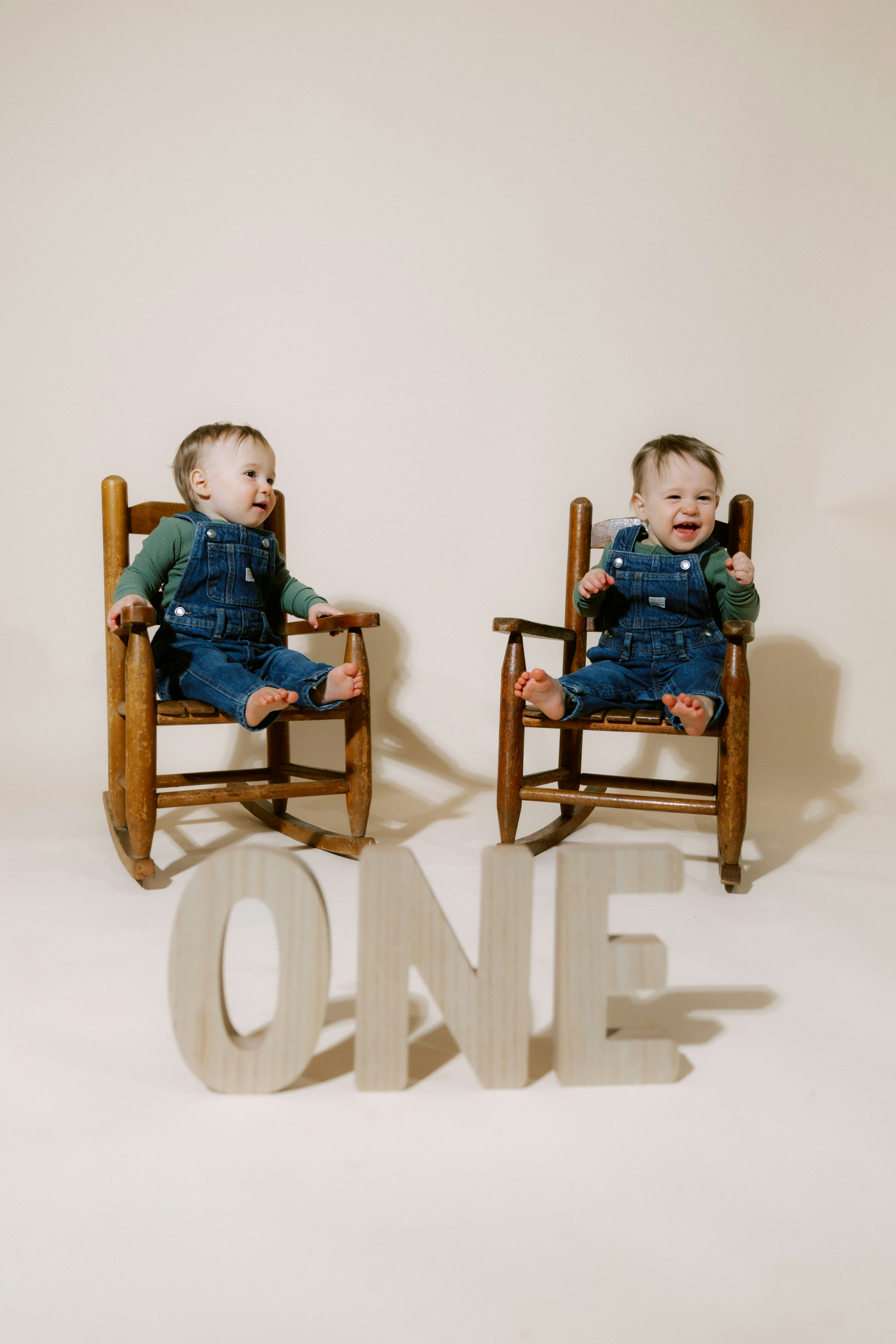 Two babies in overalls sitting in rocking chairs.