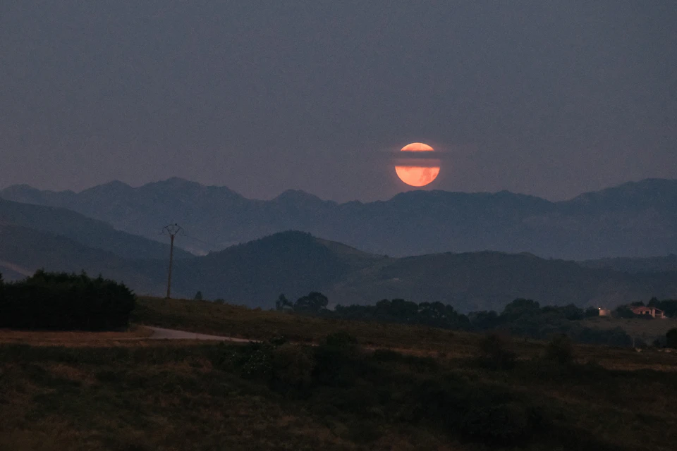 The image depicts a serene night scene with a full, radiant red moon casting its glow over a mountainous landscape. The moon is positioned centrally in the frame, illuminating the surrounding area and creating a striking contrast against the dark sky. In the foreground, there are trees and bushes that add depth to the scene. The mountains in the background rise majestically, their peaks shrouded in darkness, while the moonlight bathes them in a warm, inviting light.

The image is taken from an elevated perspective, providing a comprehensive view of the landscape below. The colors in the photo are predominantly red and blue, with the red moon standing out against the dark sky and the blue hues of the mountains adding depth to the scene.
