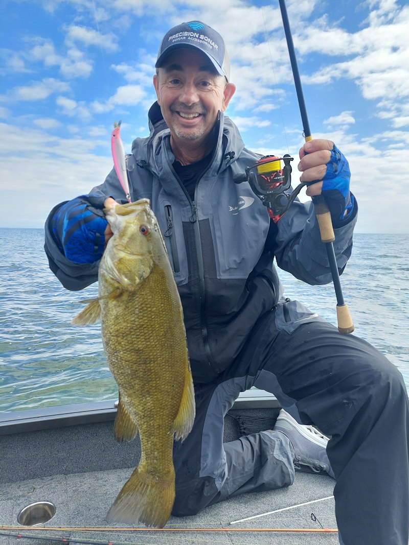Angler in a grey rain shell kneeling in a boat holding a bronze smallmouth bass with a pink jerkbait in its mouth, whitecaps behind