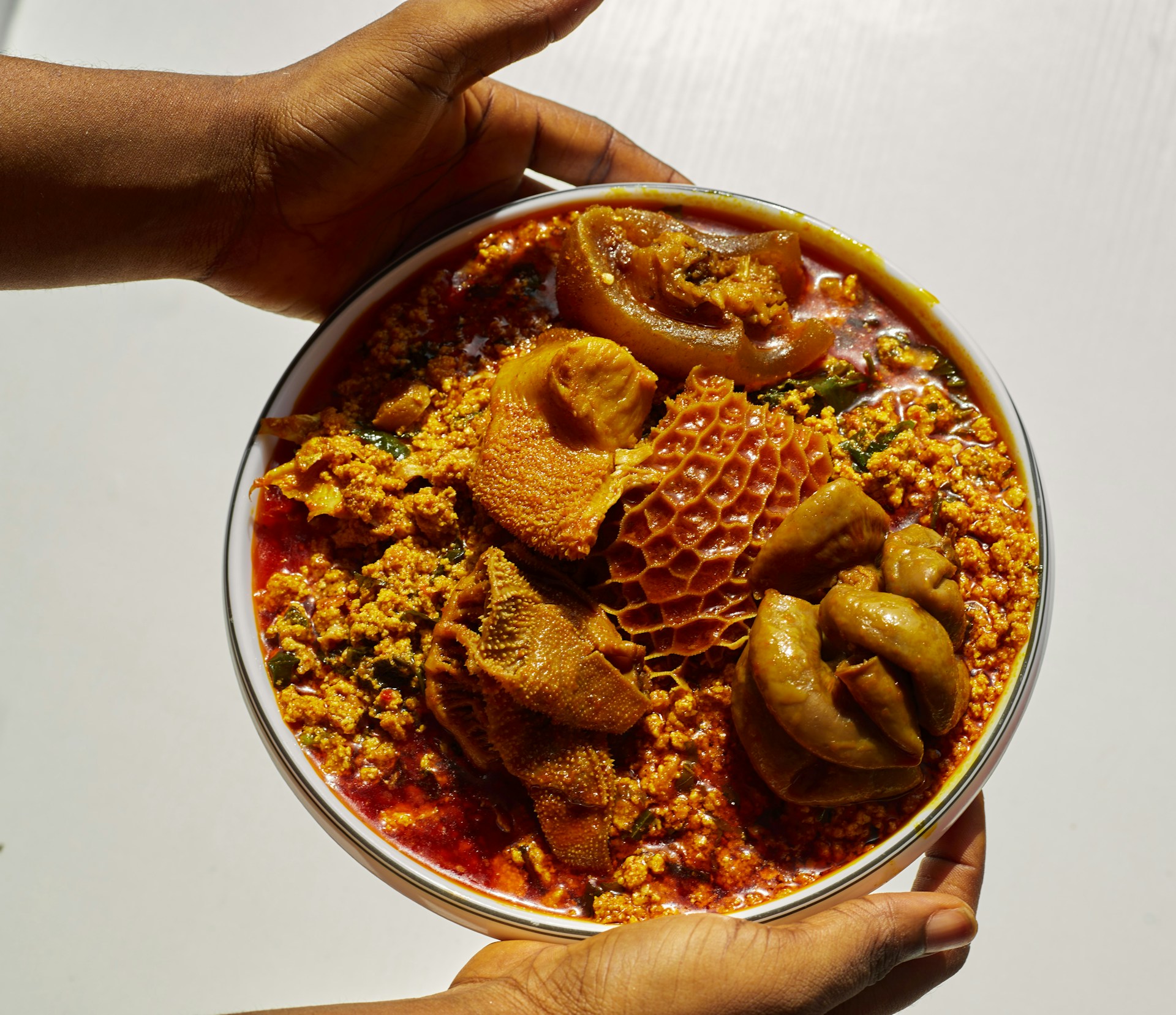 Hands holding a bowl of egusi soup with assorted meats.
