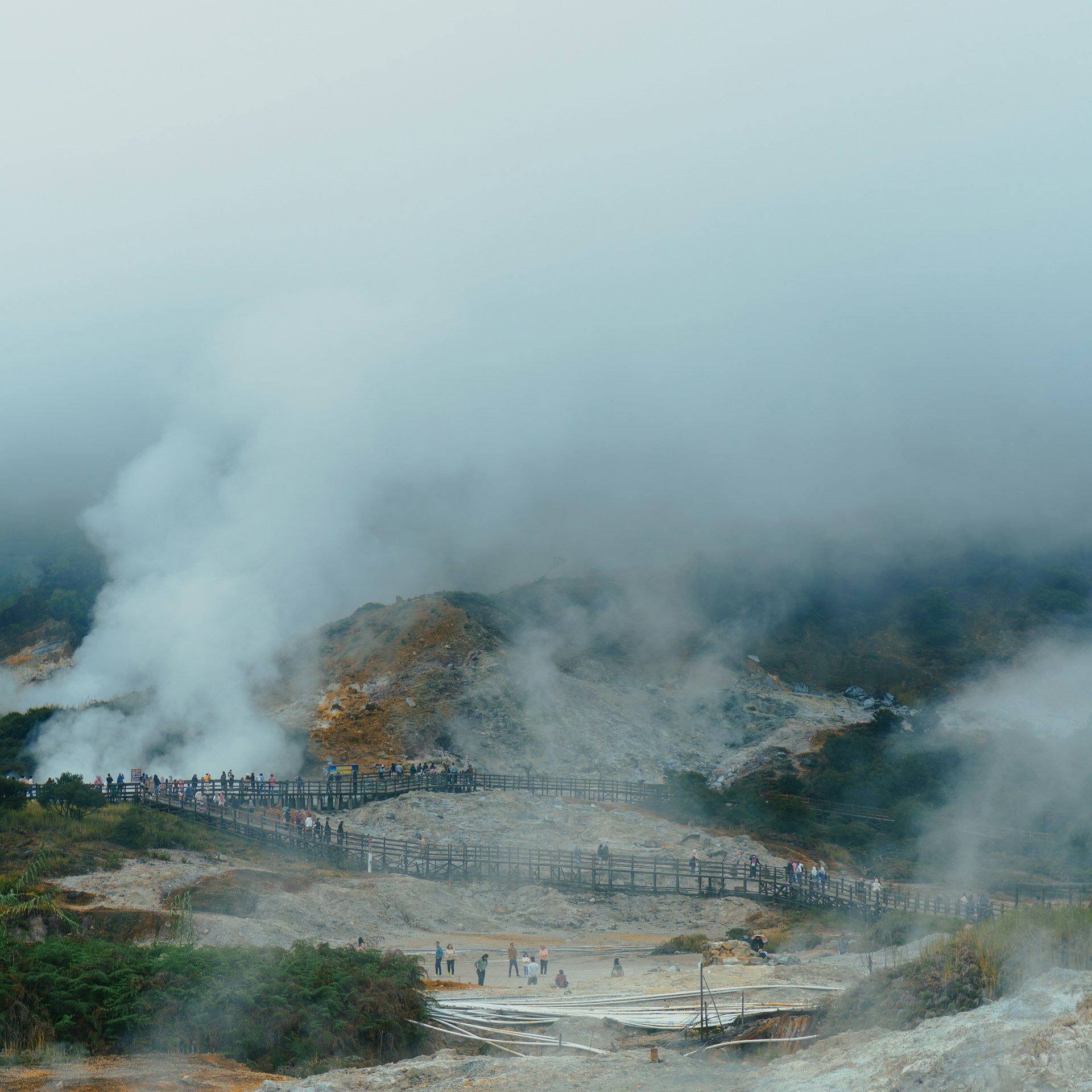 People walk on a boardwalk through steaming geothermal landscape.