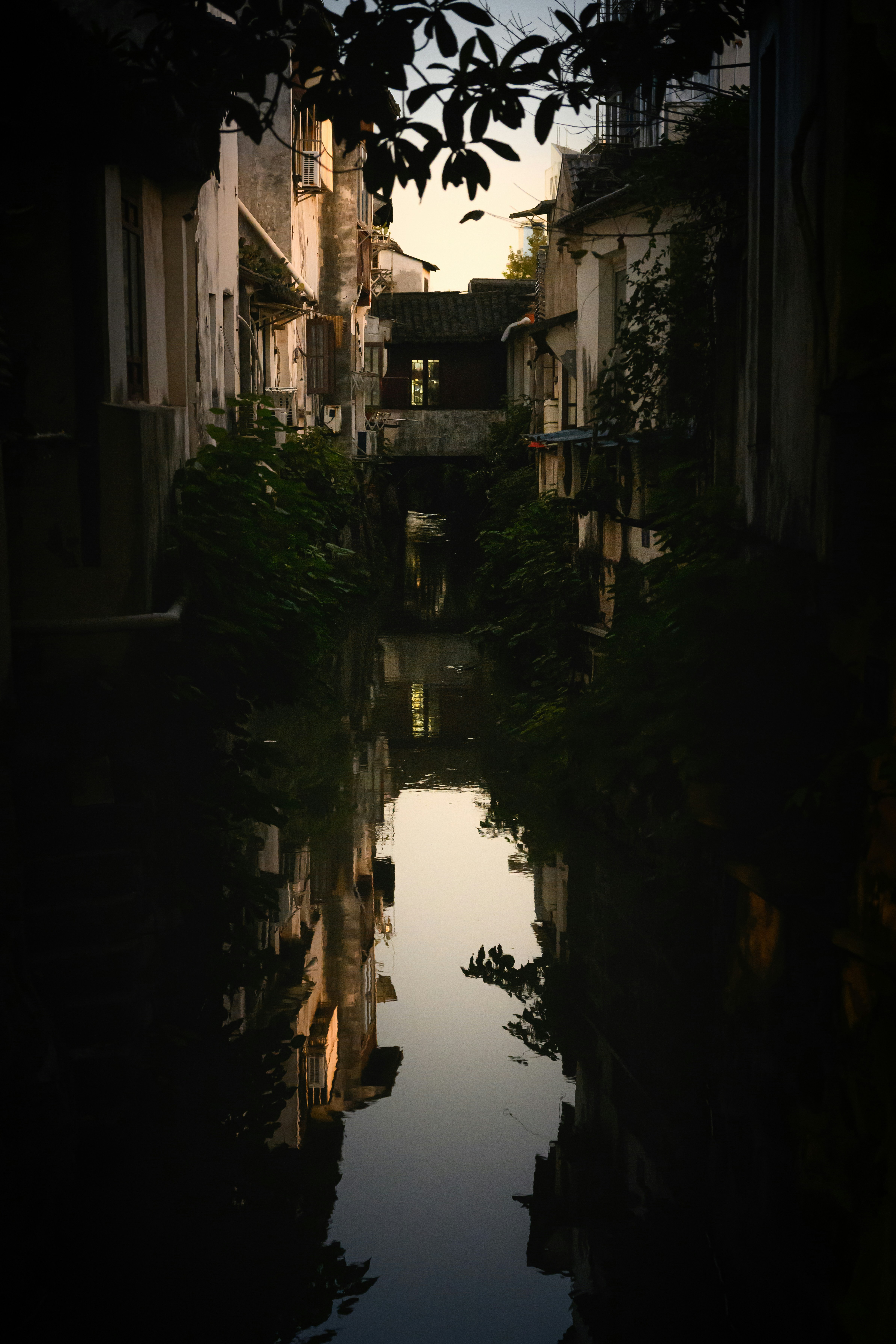 Narrow canal reflecting buildings and foliage at dusk.