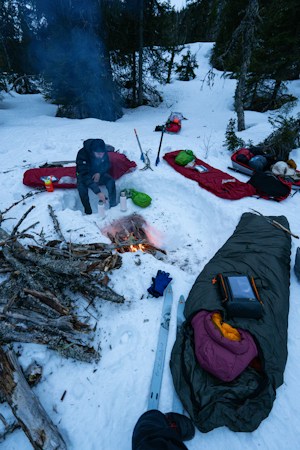 Winter camp on snow with red and dark green sleeping bags laid out on pads around a small campfire, a camper sitting on a snow bench beside water bottles