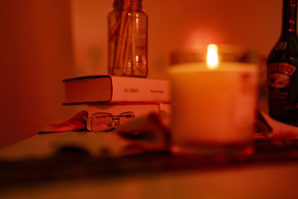 Lit candle on a desk next to stacked books and reading glasses on an open page