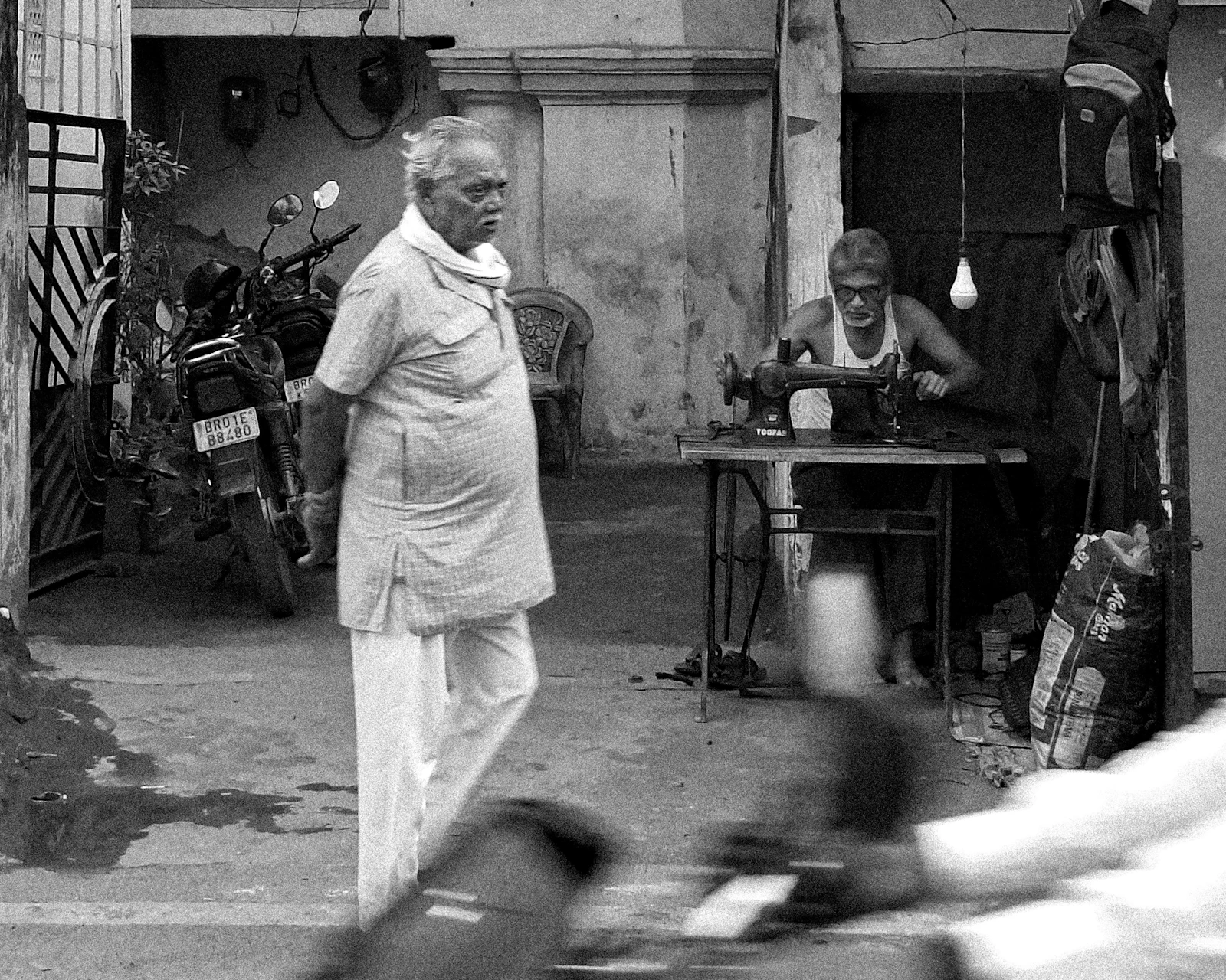 A tailor at a sewing machine as a man walks past the shop