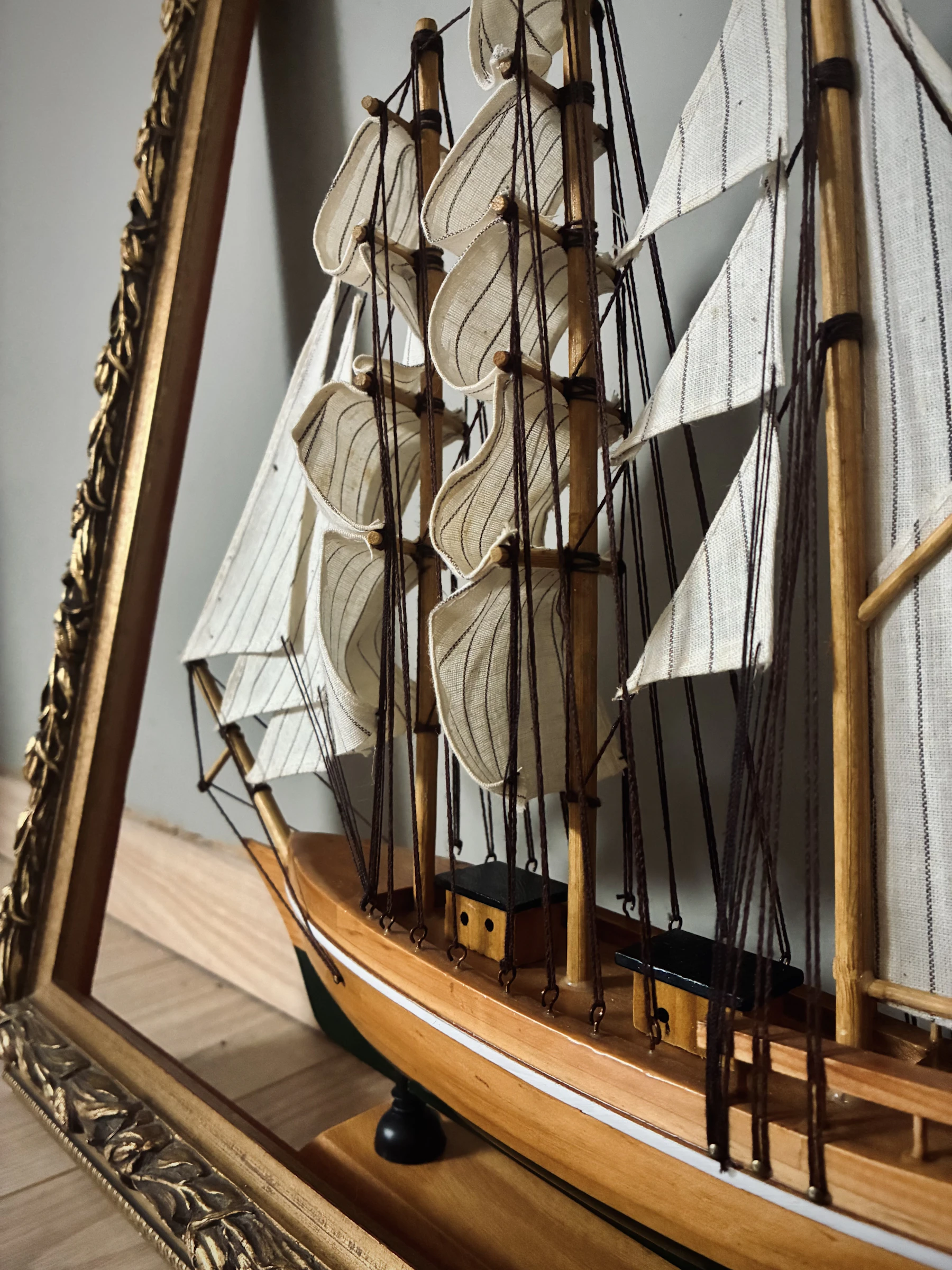 A model sailing ship with striped cloth sails and a varnished hull beside an ornate gold frame