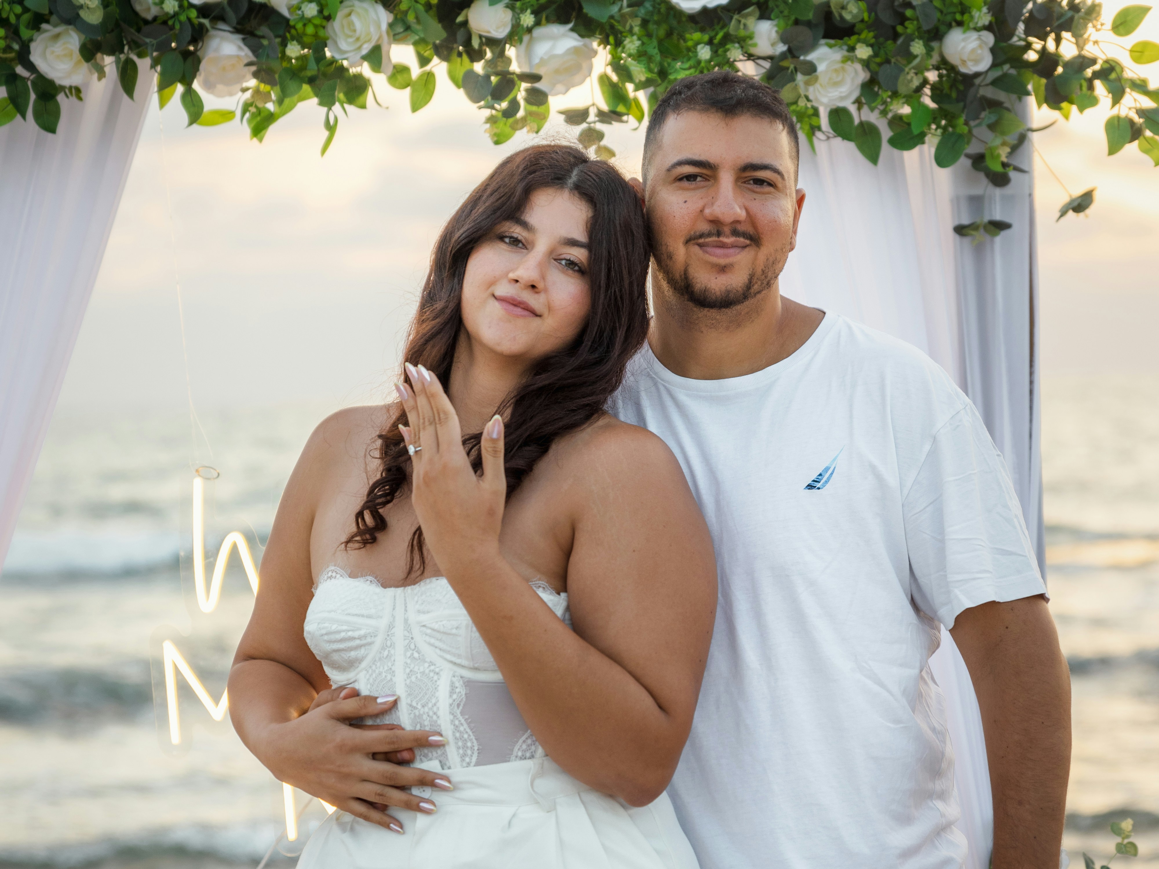A couple embraces on a beach with a wedding arch.