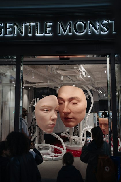 Two oversized, lifelike silicone face masks, one with eyes closed, displayed in the glass entrance of a brightly lit store