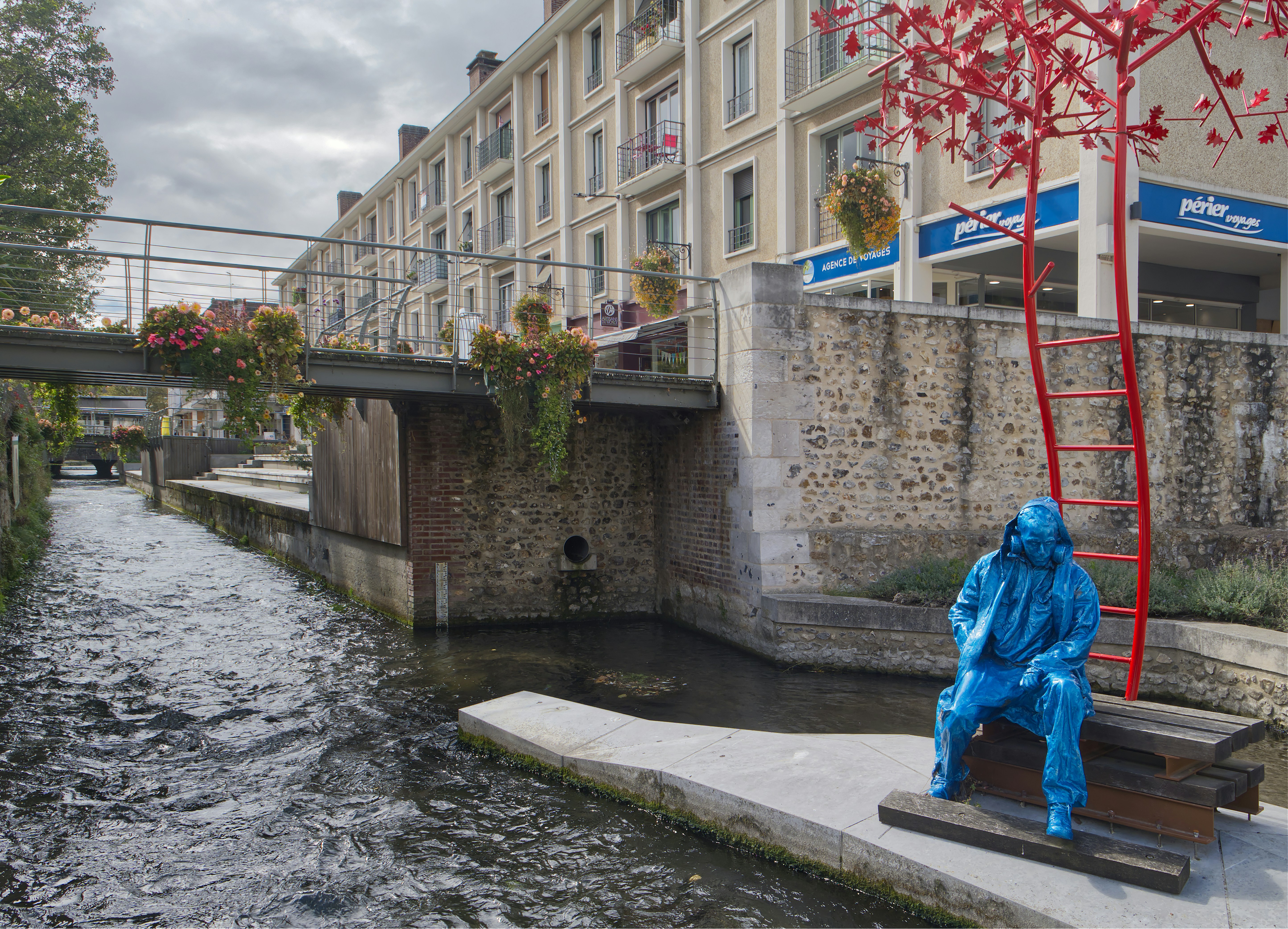 Blue statue sits by a canal with buildings behind buildings.