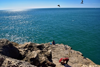 Two people sit on rocky cliff overlooking the ocean