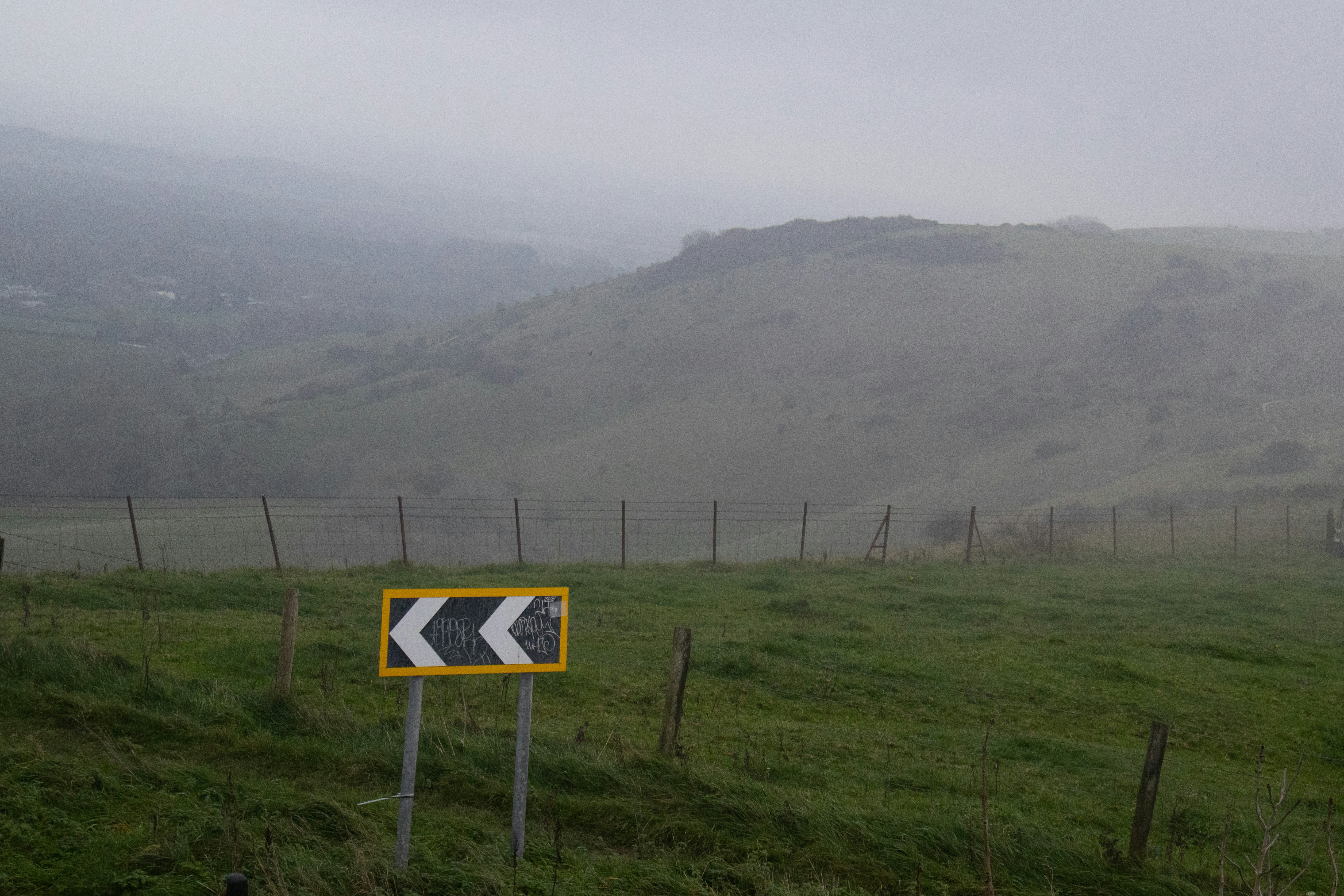 Ein Chevron-Schild auf einer nebligen Hangstraße mit Hügeln.