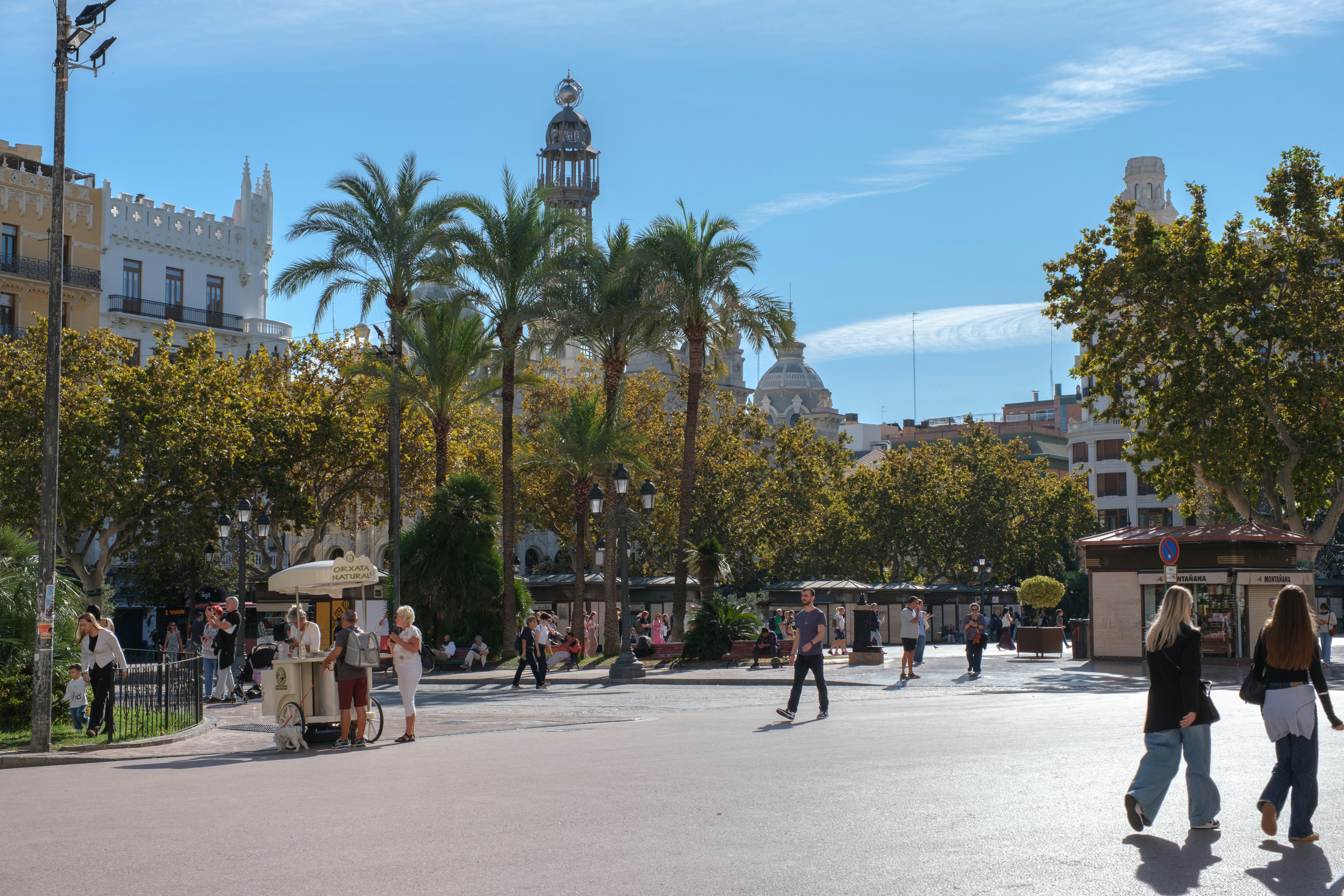 People walking in a sunny city square with palm trees.
