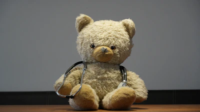 A teddy bear wearing a stethoscope sits on floor.