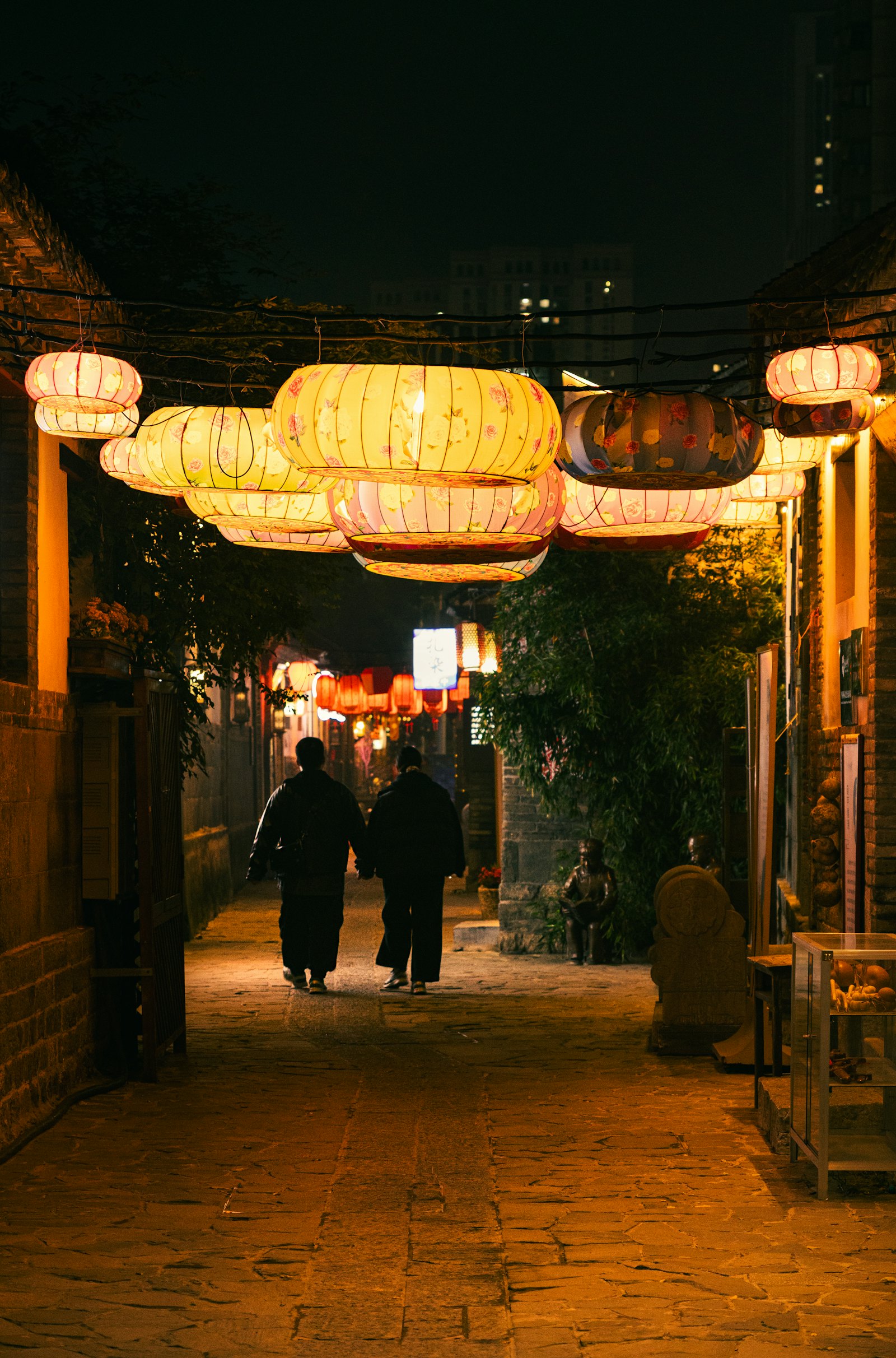 Two visitors seen from behind walking a lantern lit old town lane at night