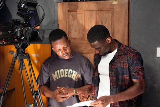 Two men looking at a script by a camera.