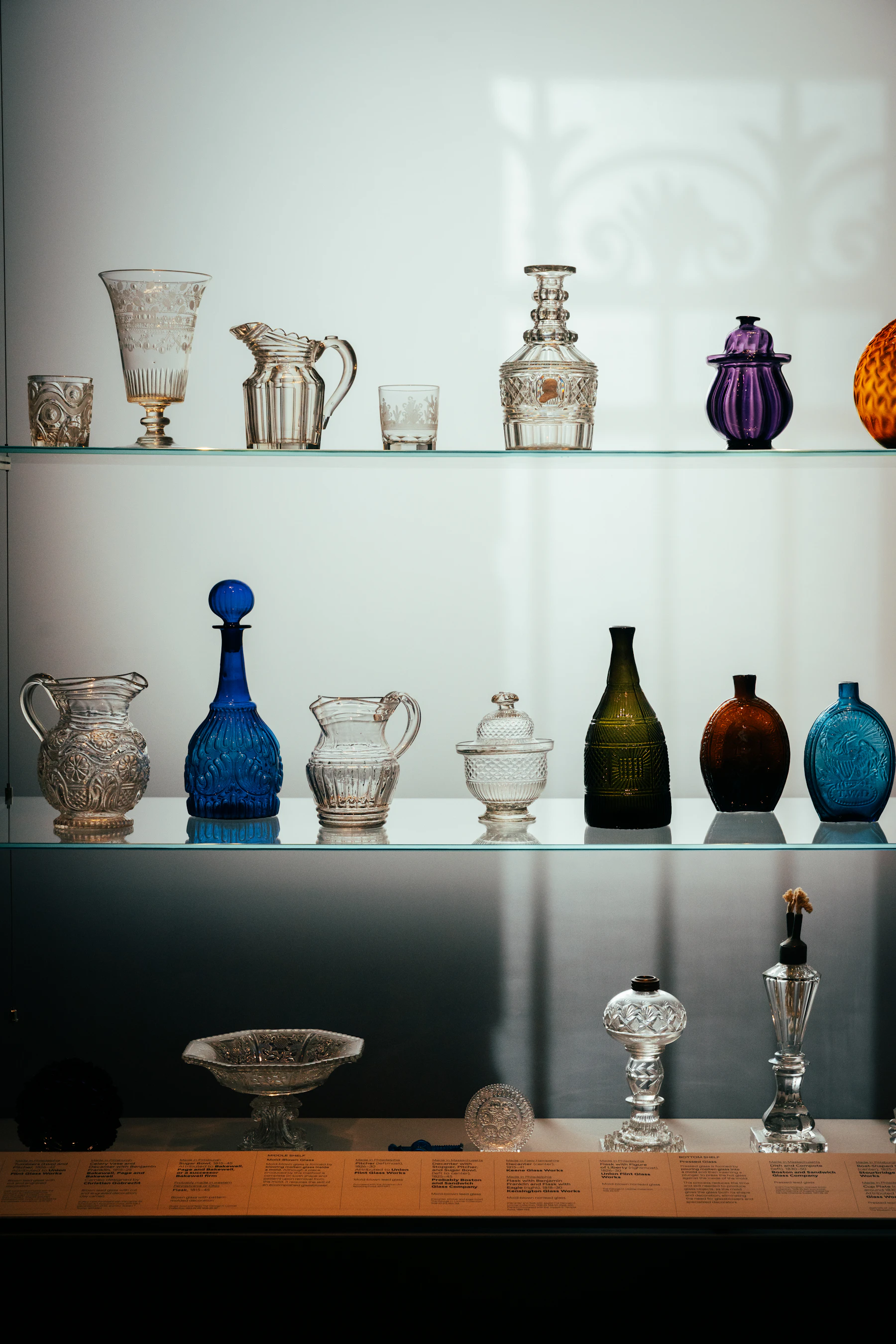 A museum case with pressed and colored glass pitchers, flasks and bowls on glass shelves