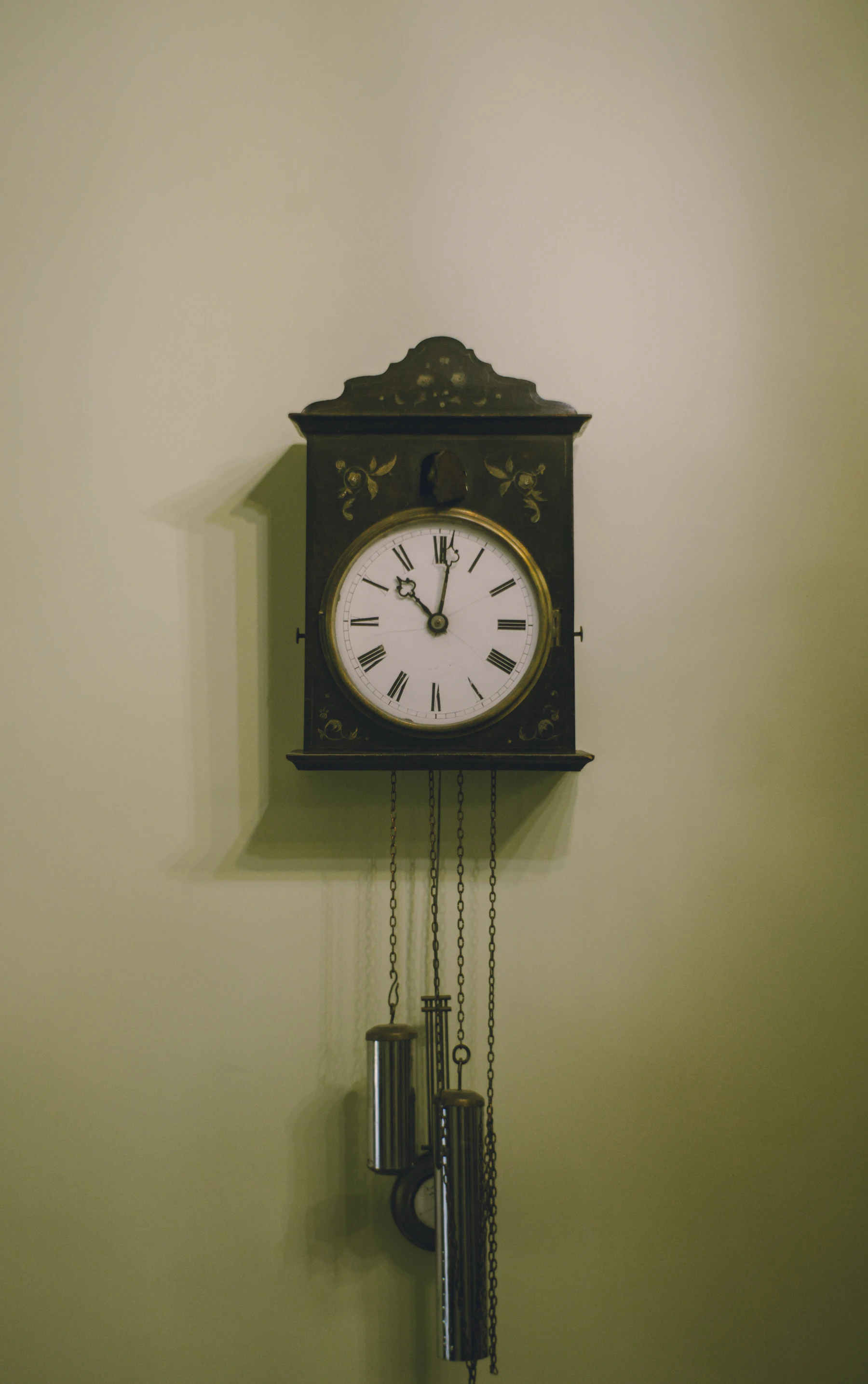 A dark painted wall clock with a white dial and two weights hanging below it on chains