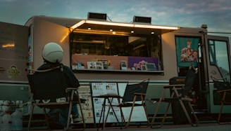 Person sits outside a brightly lit food truck at dusk.