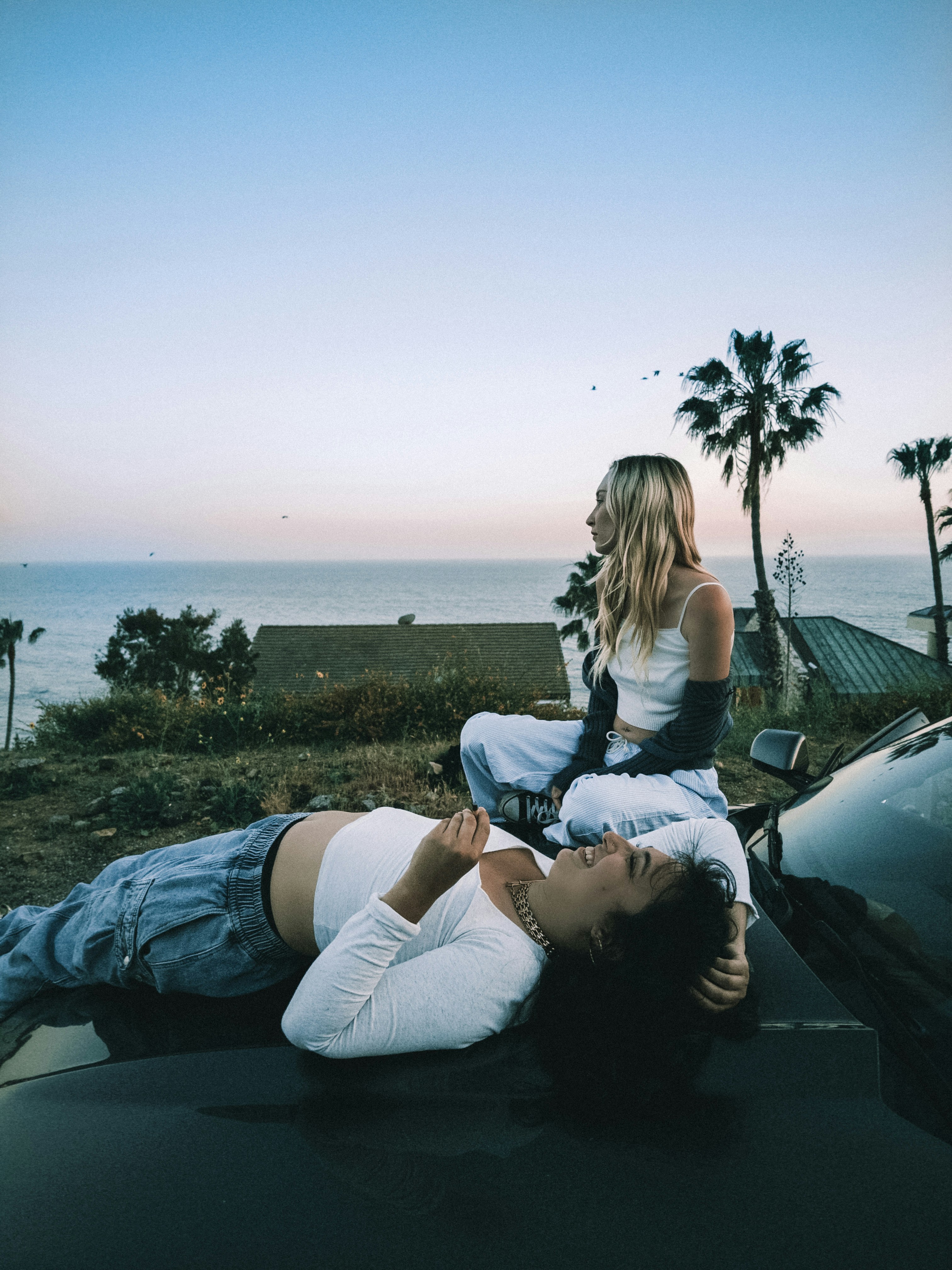 Two friends relax on a car overlooking the ocean.