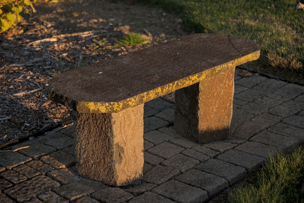Natural stone bench positioned along a garden walkway, dappled in afternoon sunlight with layered greenery behind