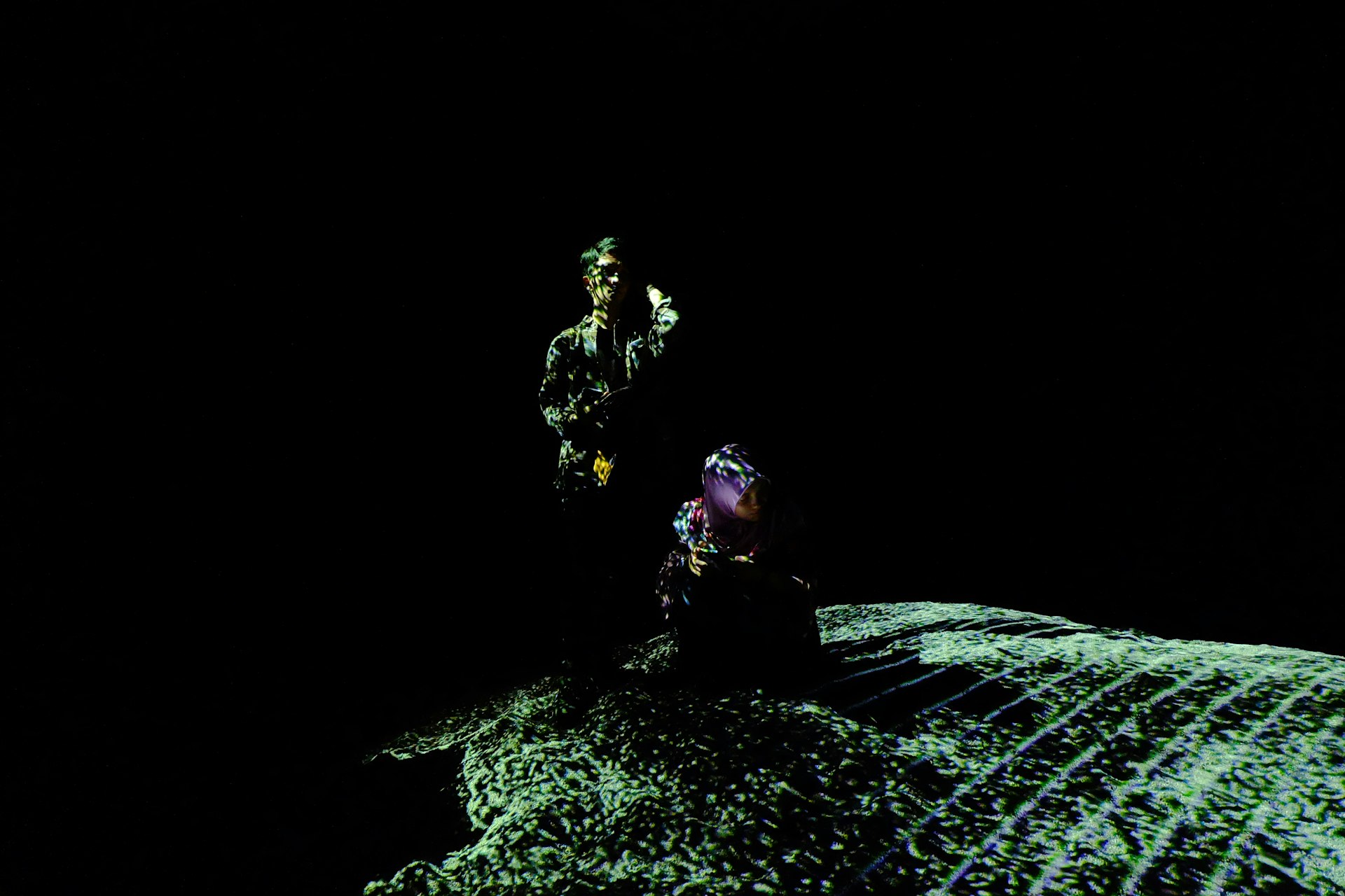 Two figures stand on a textured surface in darkness.