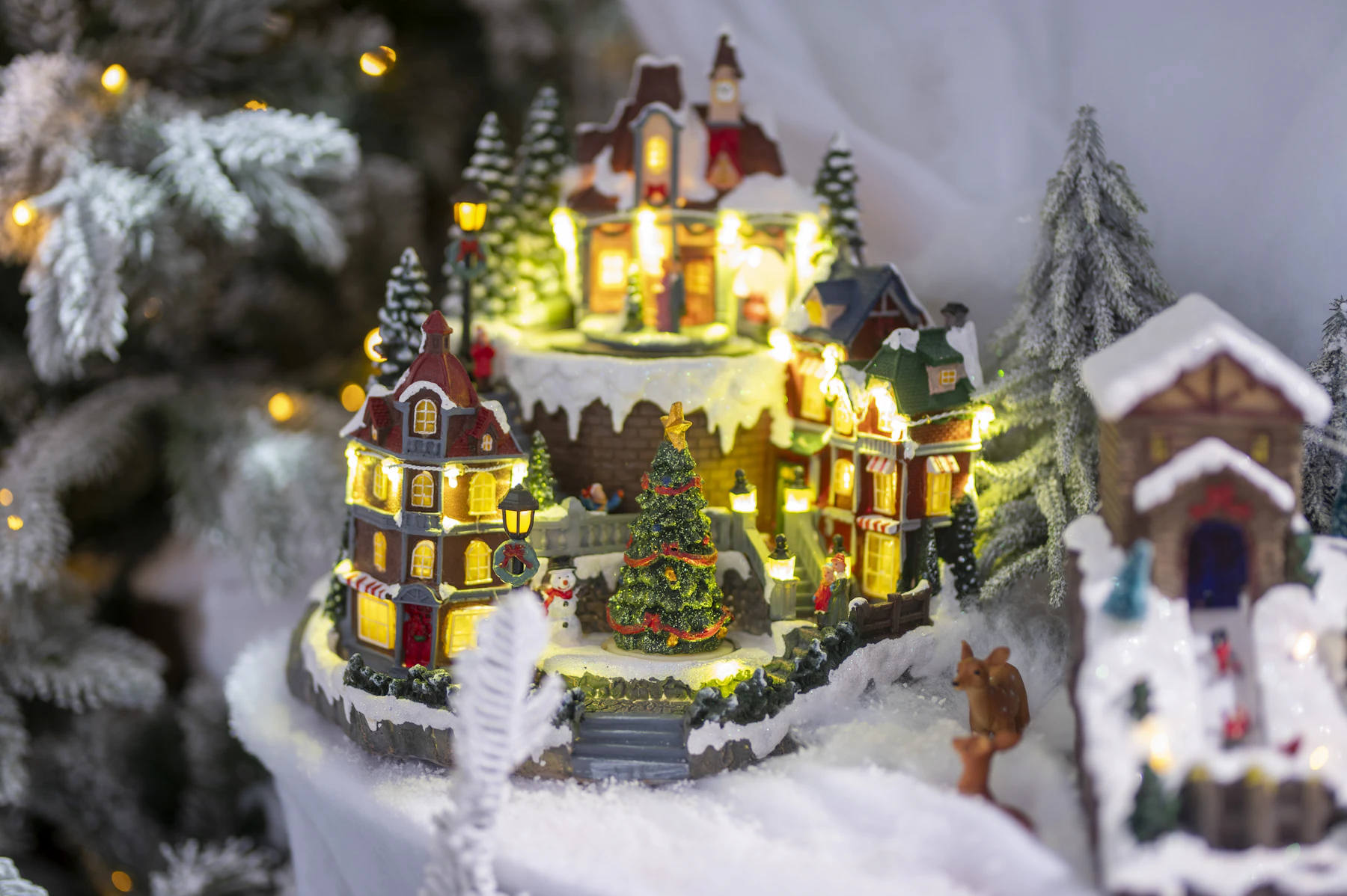 A lit resin village piece with shops on two snowy levels, a small tree in its square, beside a flocked Christmas tree