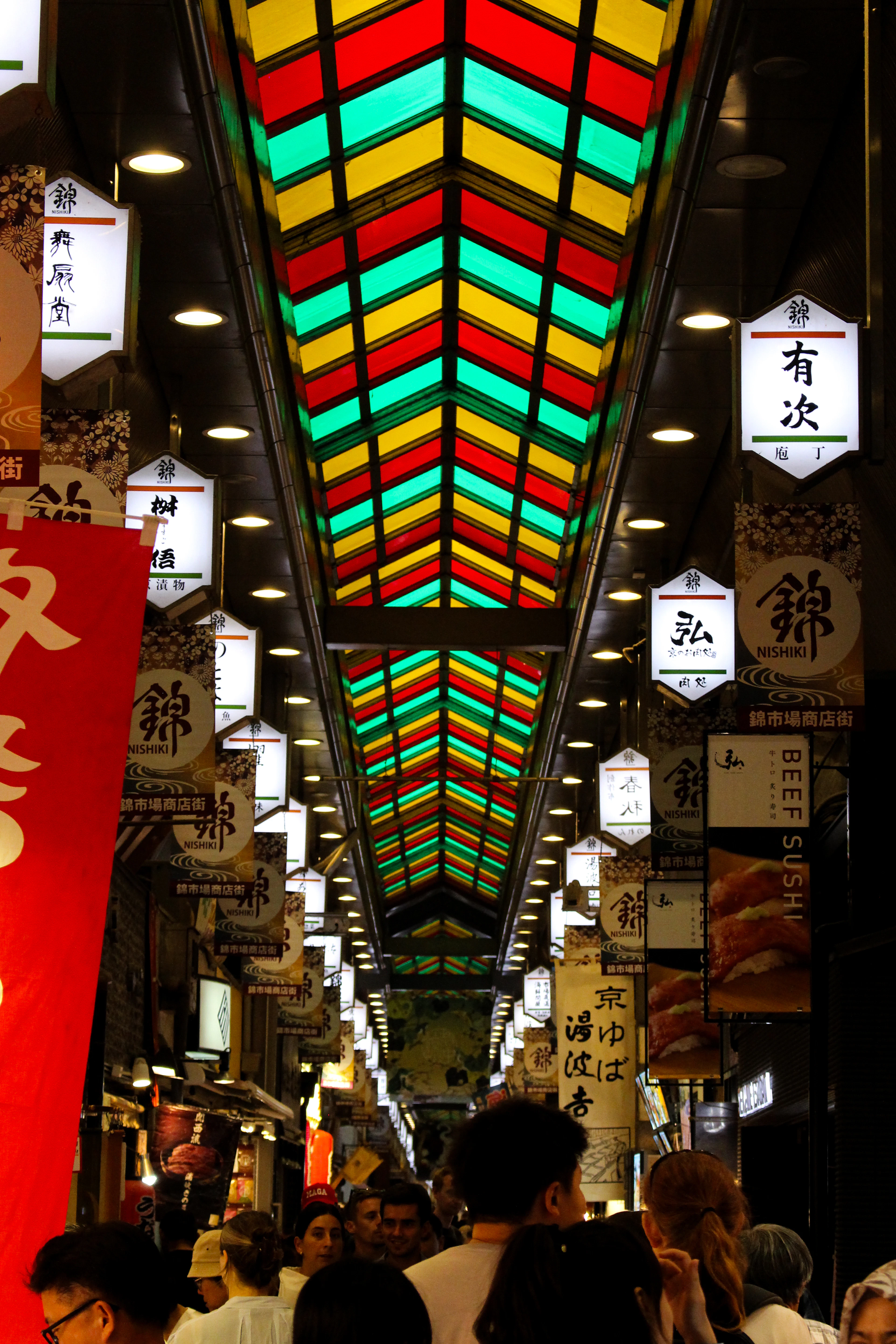 Shoppers moving beneath the red, green and yellow glass arcade roof of Kyoto's Nishiki Market, hanging banners marking each stall down the covered lane.