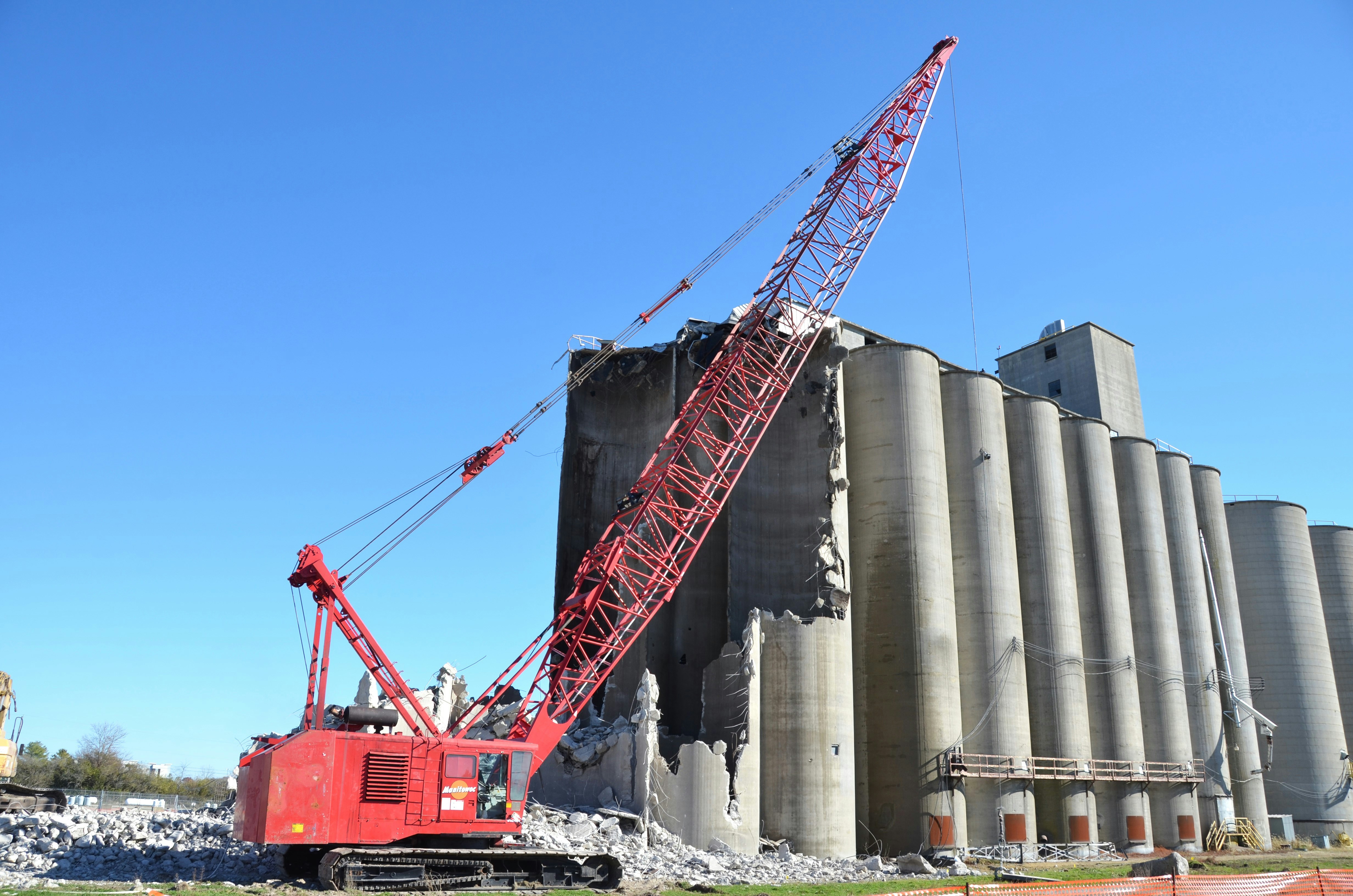 Crane demolishing concrete silos against clear blue sky.
