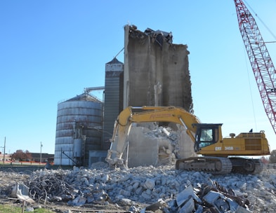 Excavator demolishes concrete silos under clear blue sky.