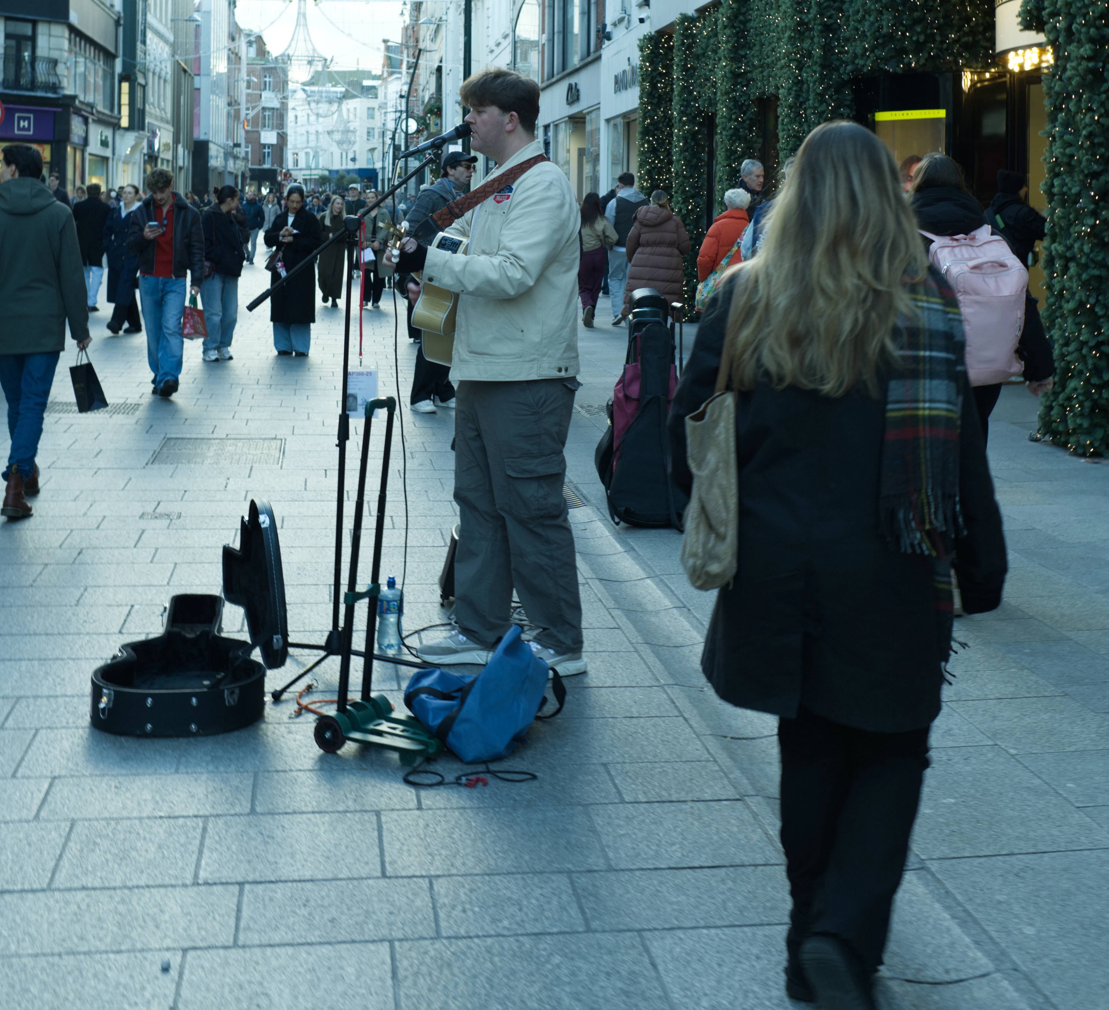 A busker singing into a microphone with an acoustic guitar on Grafton Street in Dublin, his open case on the paving in front of him, shoppers streaming past under shopfronts hung with garlands of fairy lights