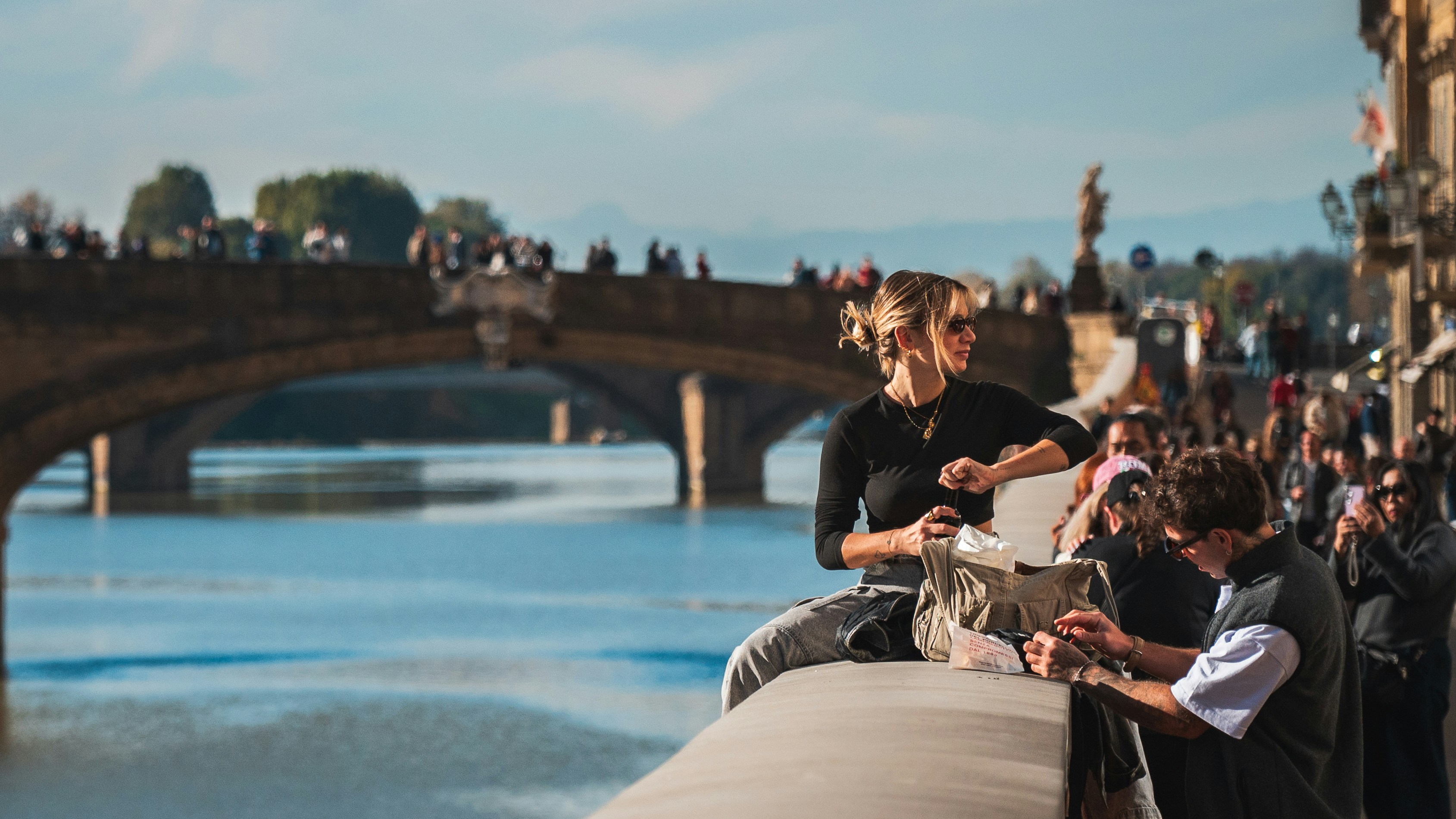Two people sitting by a river with a bridge.