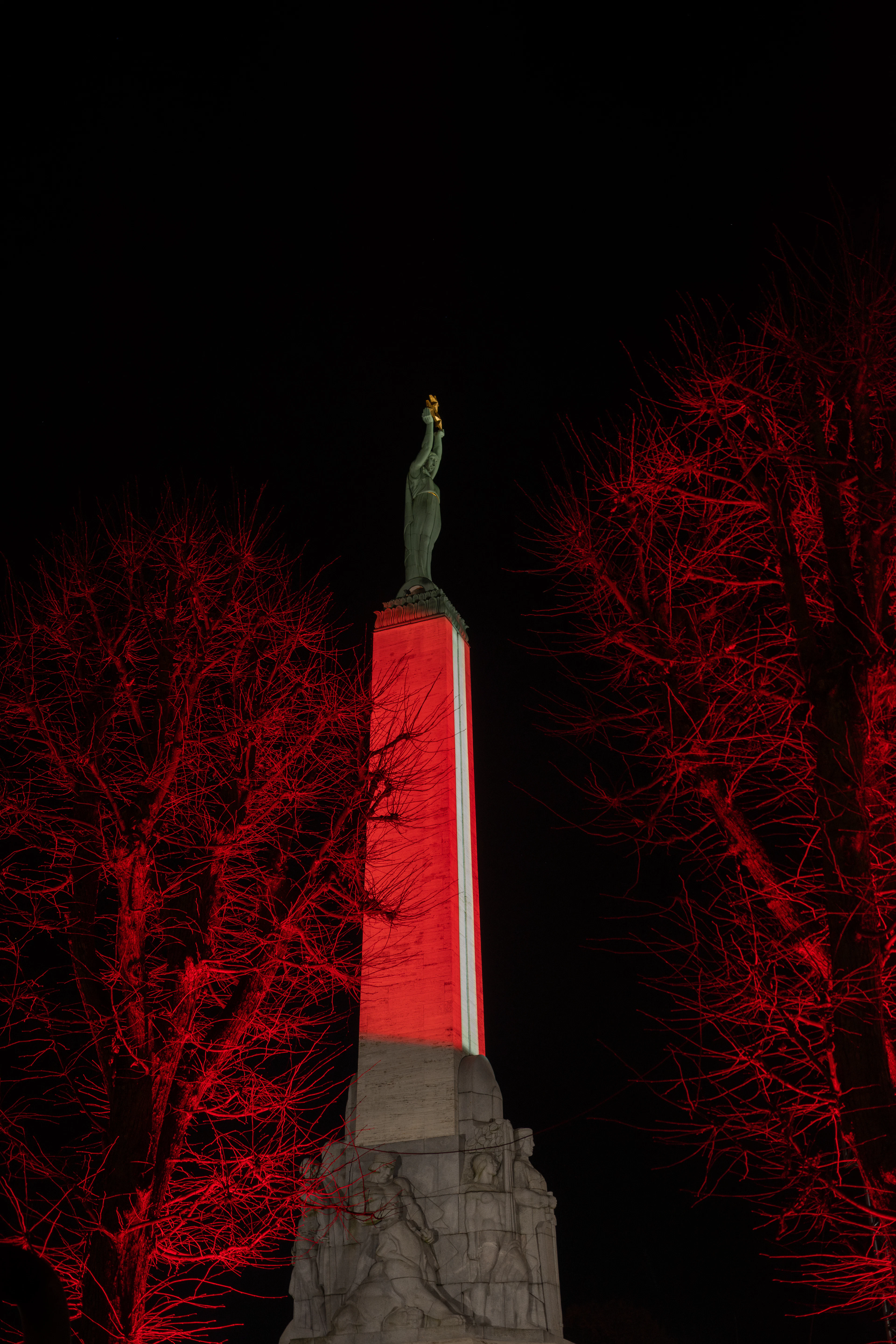 The Freedom Monument in Riga floodlit in the red-white-red of the Latvian flag at night, the bronze figure of Milda raising her three gilded stars above the column, framed by bare trees washed in red light.