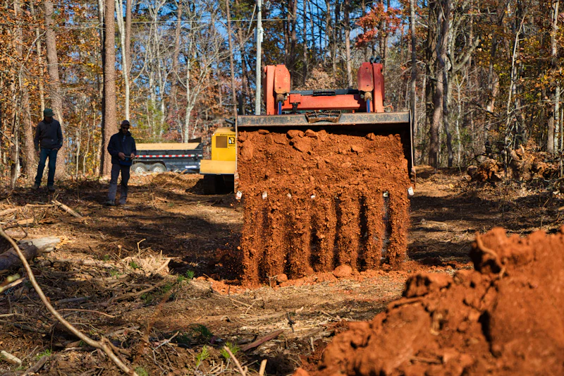 Two workers watching an excavator dig into red clay on a wooded site