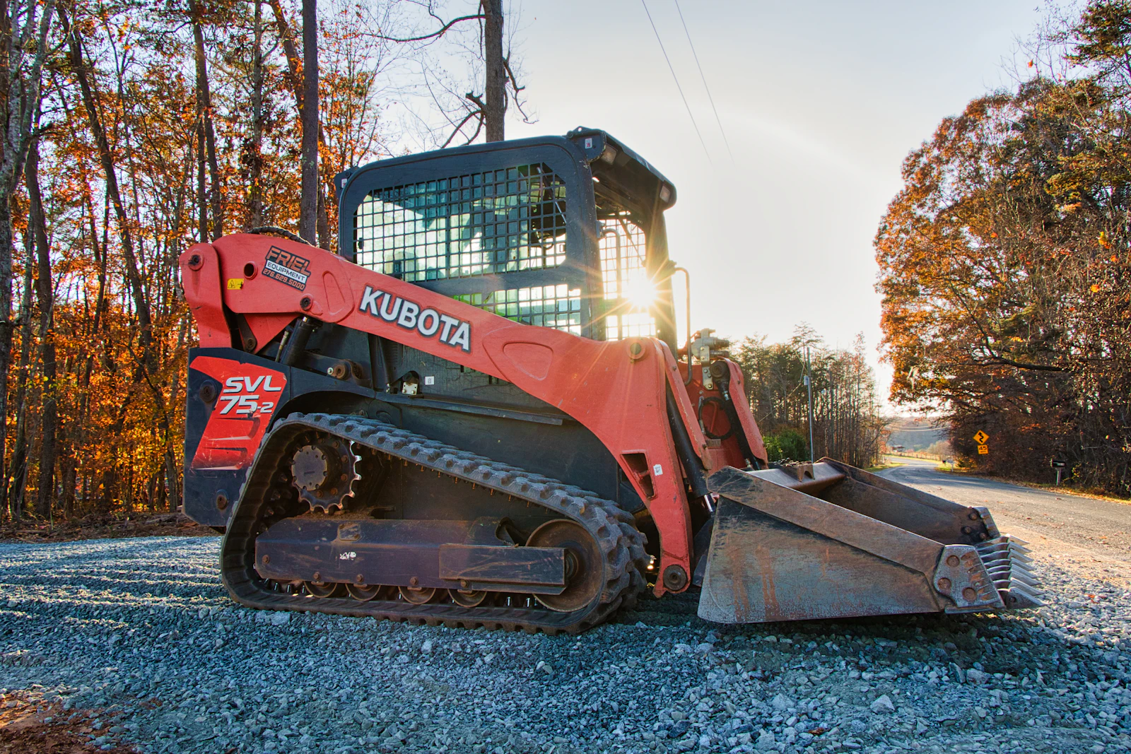 A compact track loader working an access lane at the edge of a wooded lot