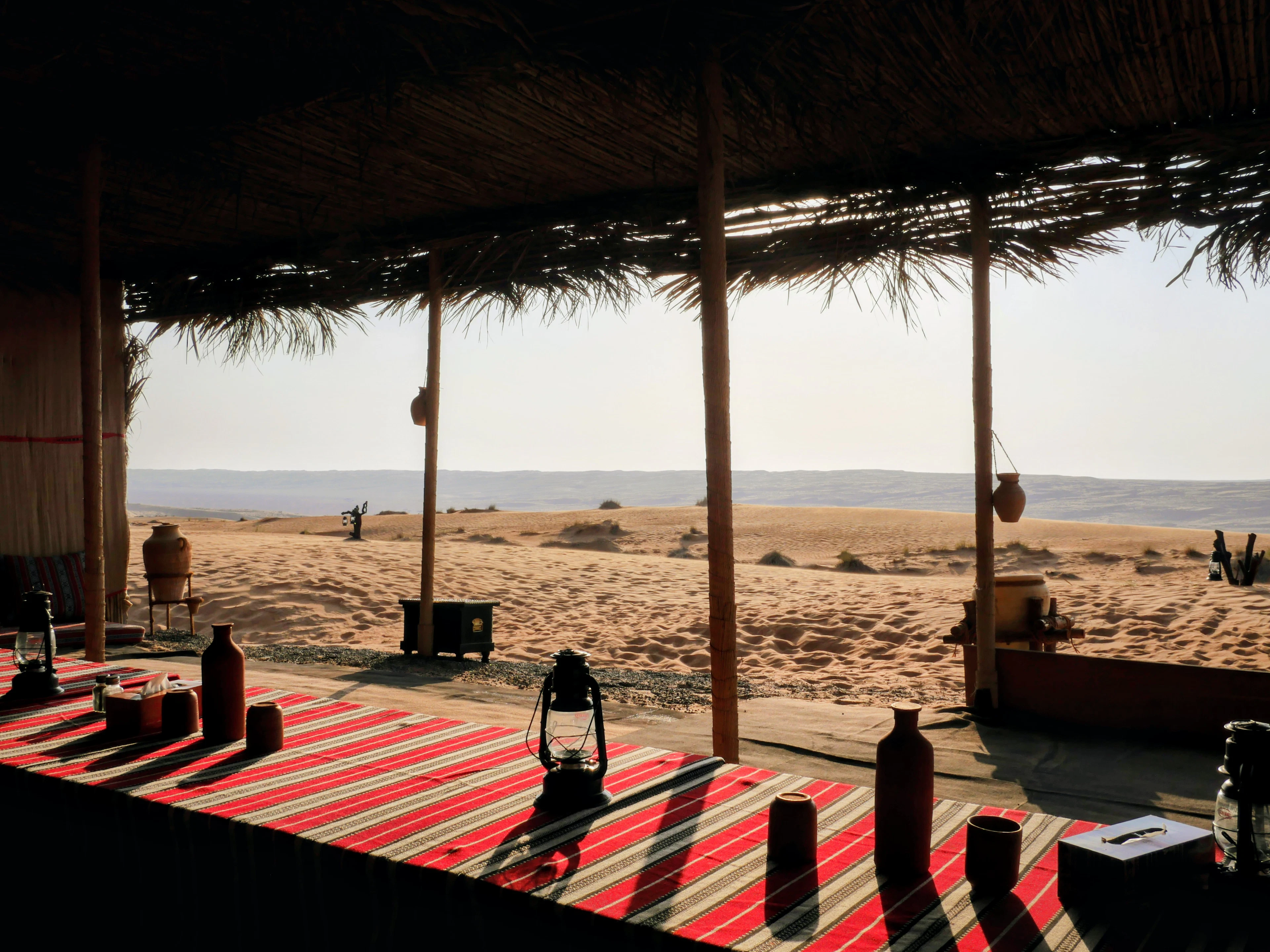 A shaded seating area with red rugs and lanterns at a desert camp, looking out to the dunes, Oman