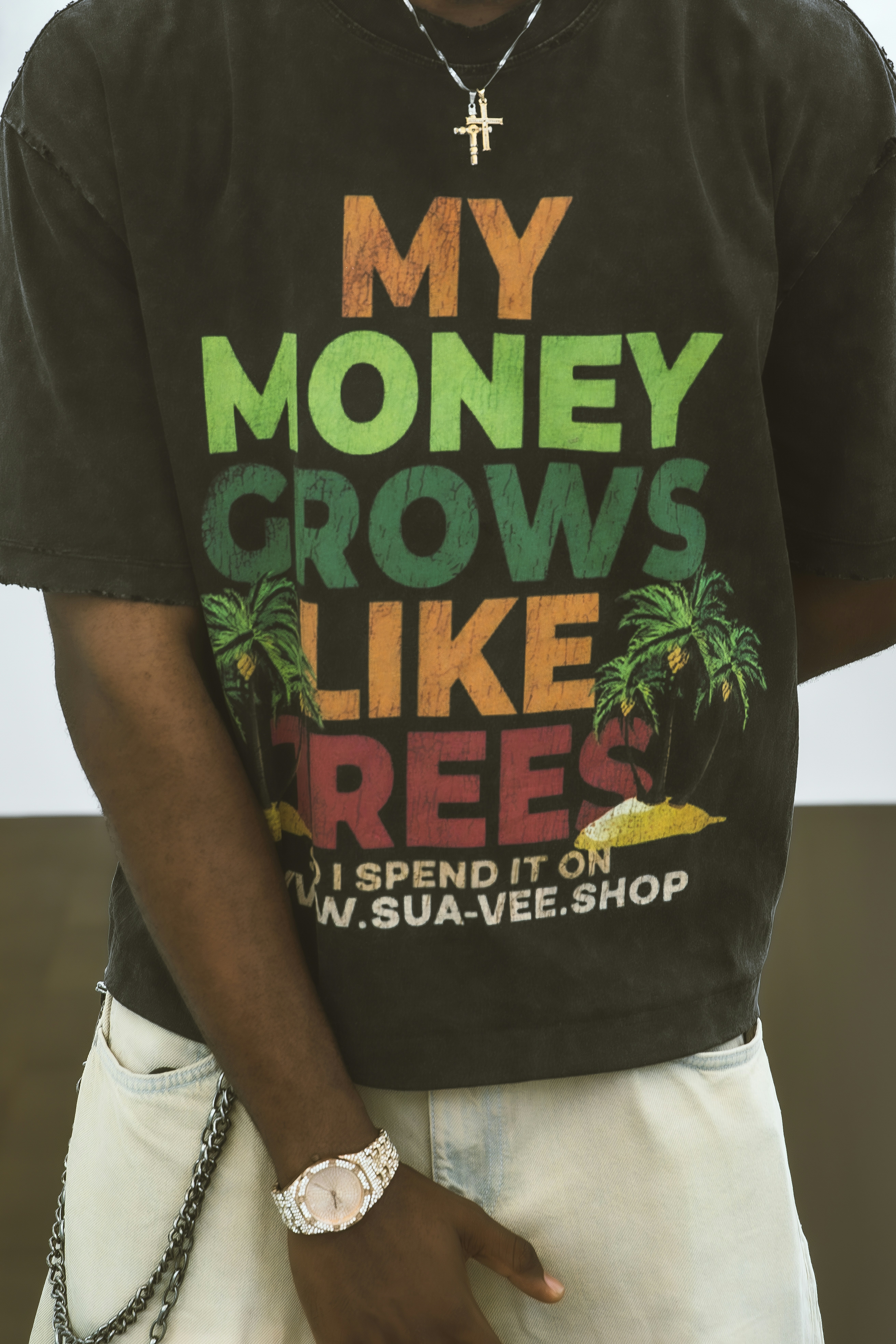 A close-up view of a graphic streetwear tee with bold, colorful lettering and palm tree illustrations. The shot captures the texture of the shirt, layered jewelry, and casual denim, creating a clean urban fashion detail.