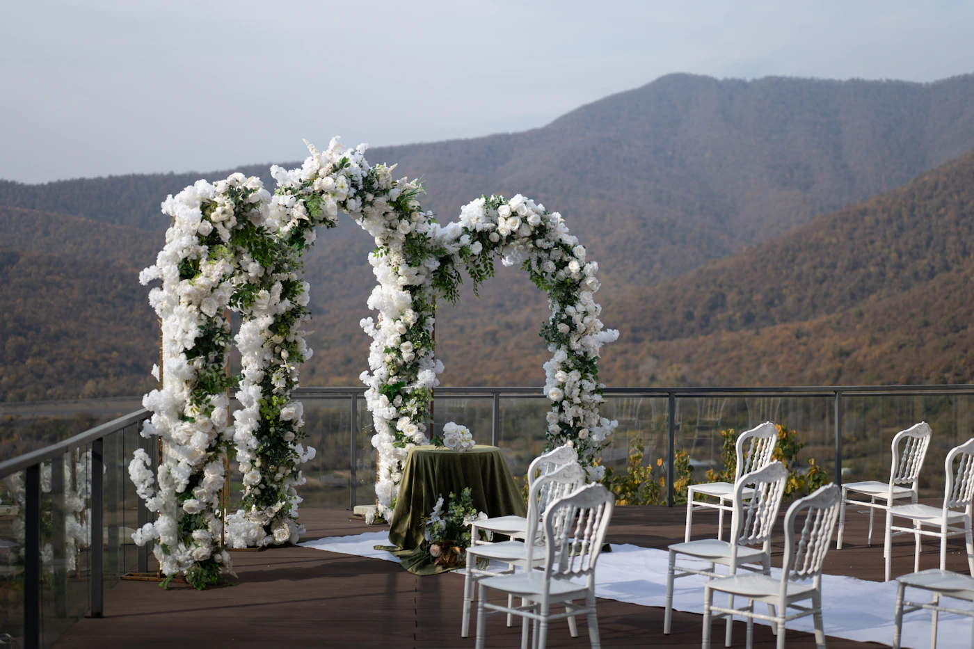 An outdoor wedding ceremony setup with a large floral arch decorated with white flowers and greenery and rows of wooden chairs arranged facing the arch with dramatic mountains rising in the background