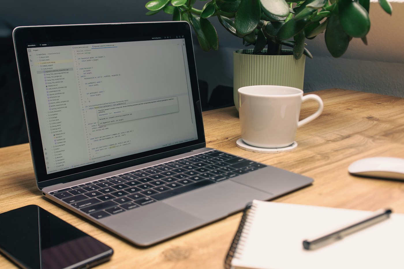 A laptop displaying code beside a smartphone, notebook, coffee mug, and potted plant on a wooden desk