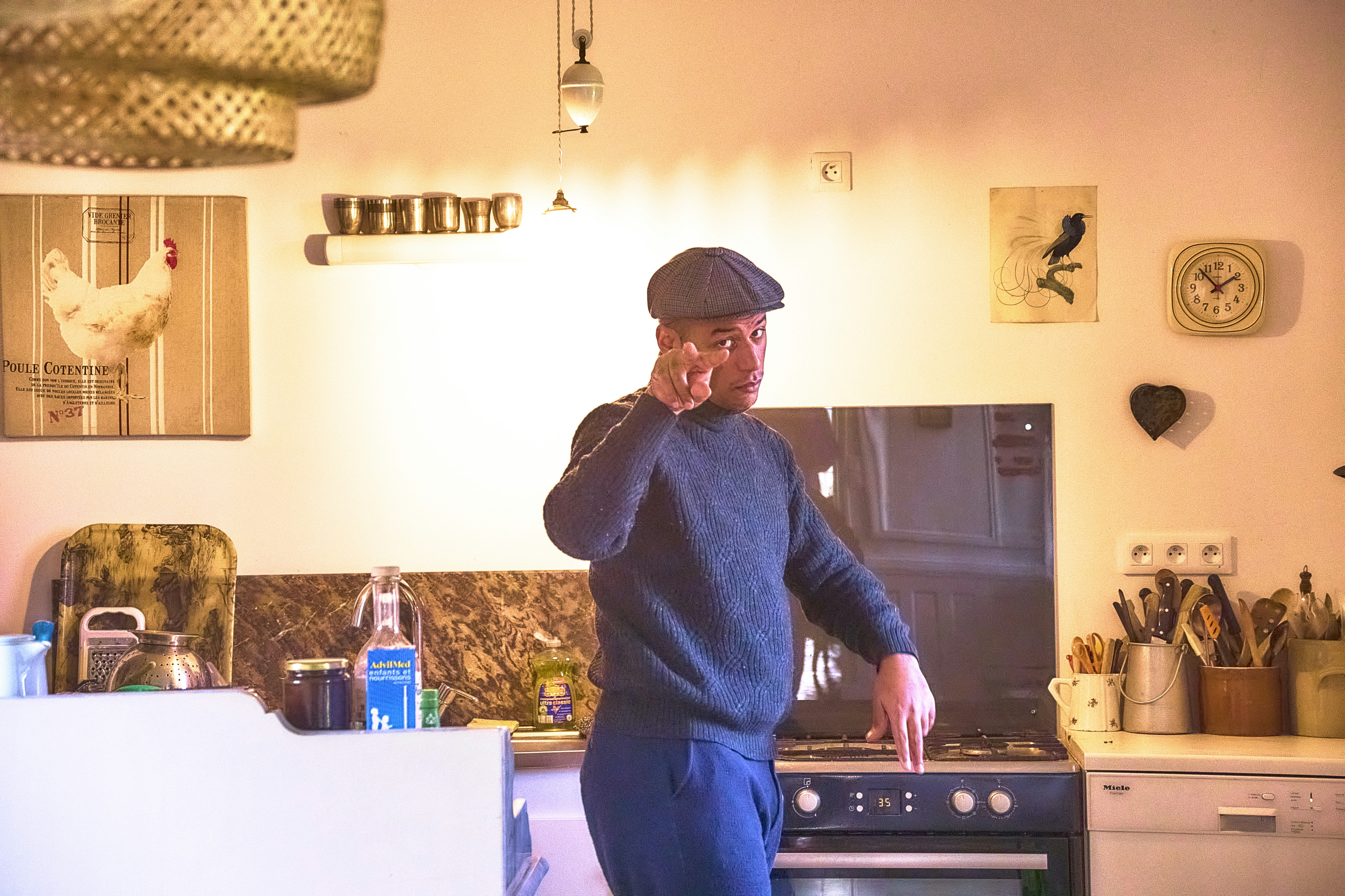 Man in flat cap talking on phone in kitchen.